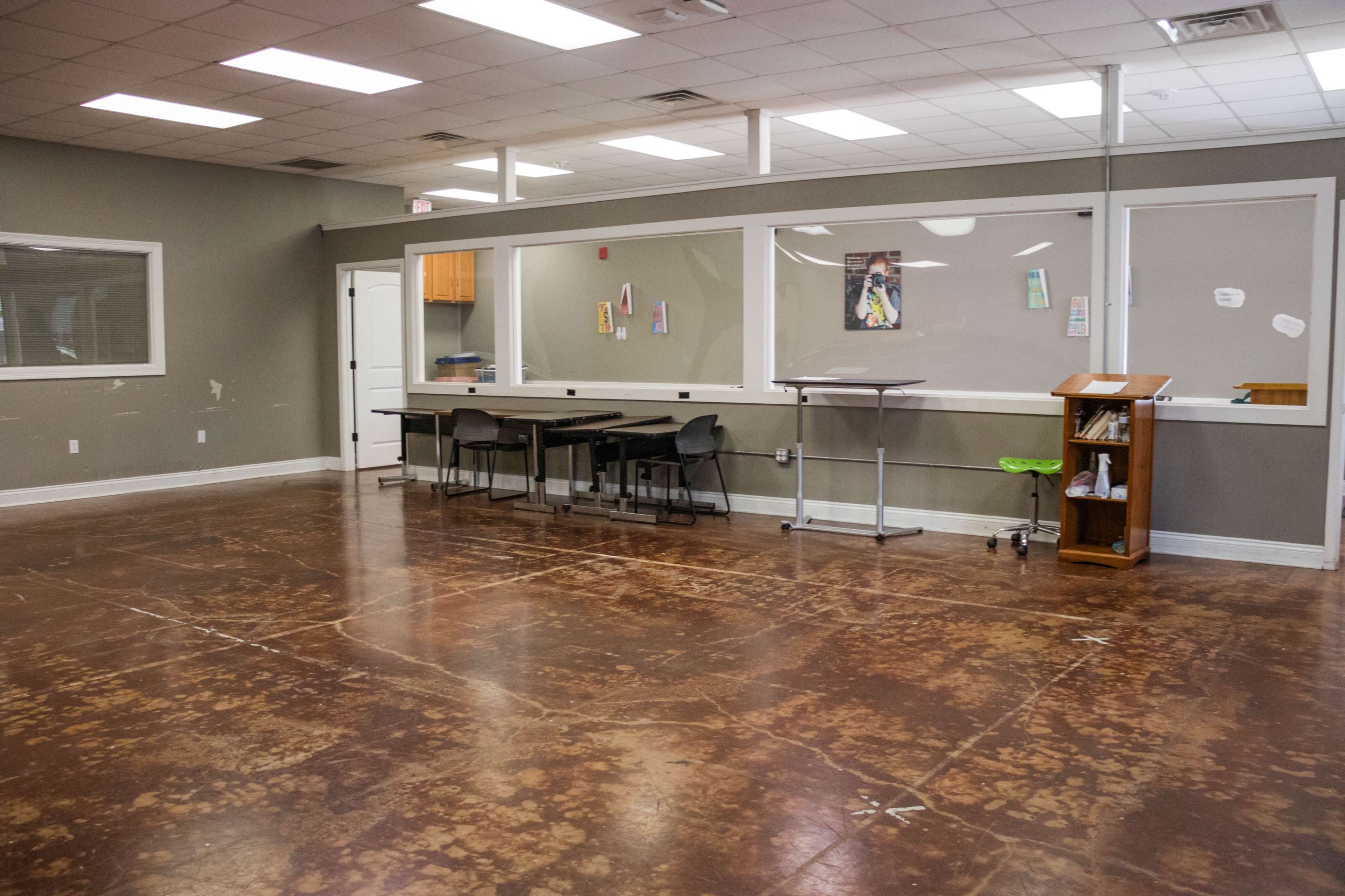 The image shows an empty classroom with polished concrete flooring, a large windowed area, and a few tables and chairs arranged in a sparse layout.
