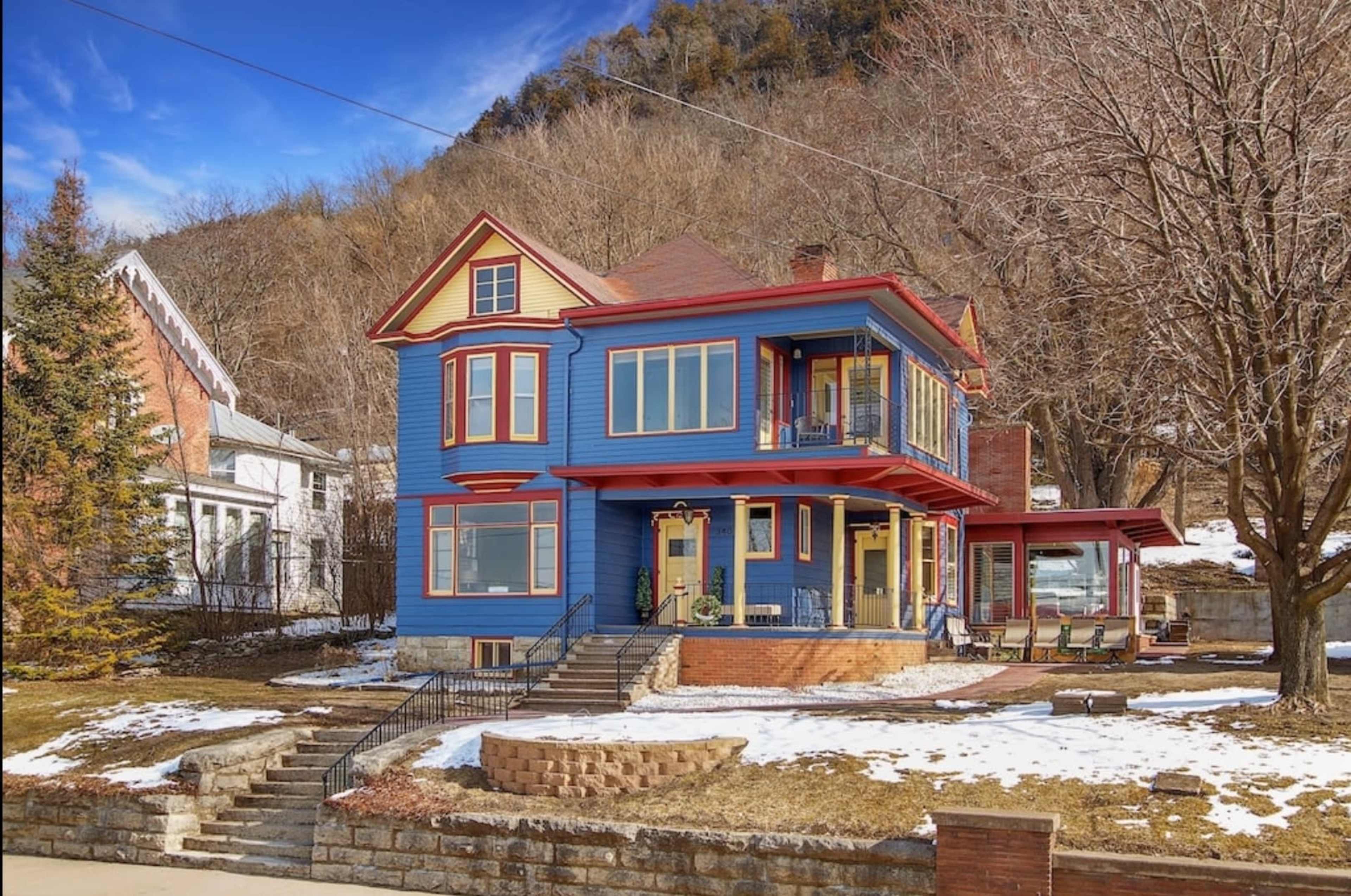 The image shows a large blue house with a red trim, featuring multiple balconies and surrounded by a snowy landscape and bare trees.