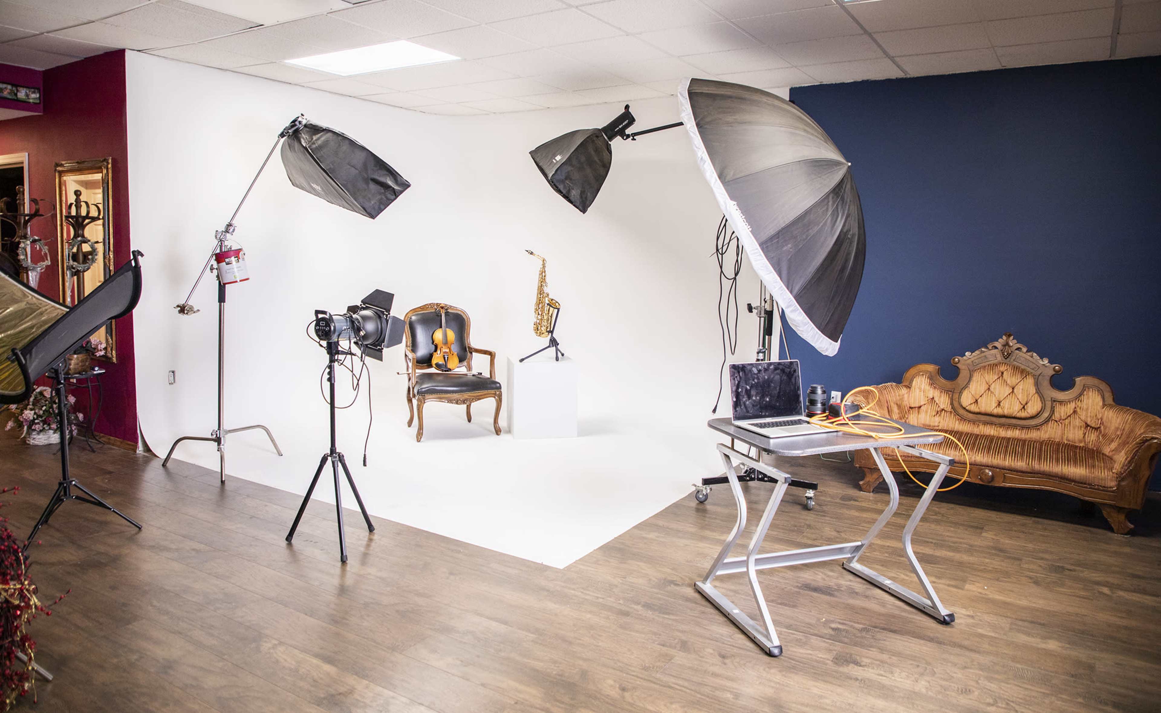 A photography studio setup with softbox lights, a camera on a tripod, a wooden chair, a saxophone displayed on a pedestal, and a laptop on a table.