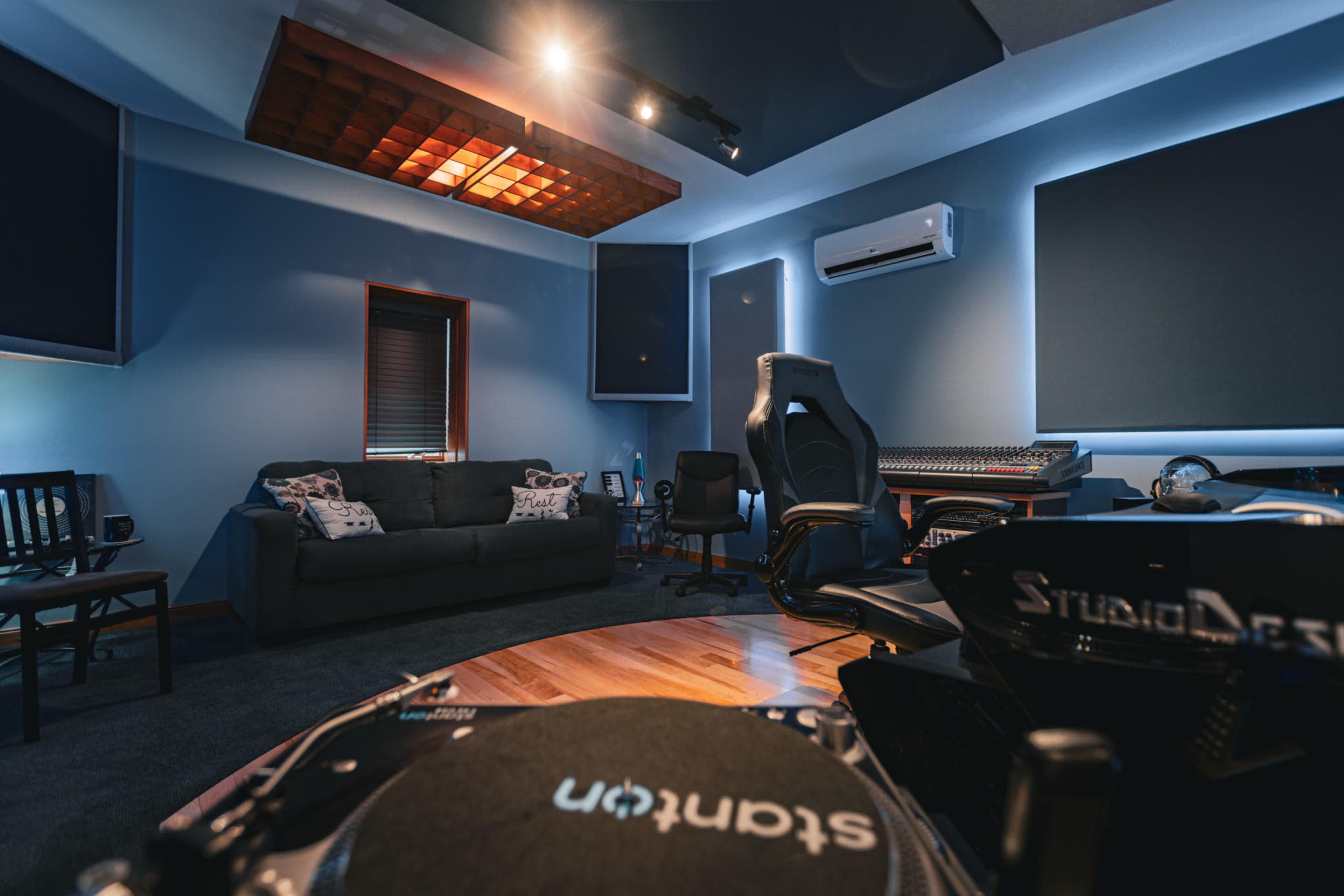 The image shows a music studio with a couch, office chair, mixing console, and soundproof panels on the walls.