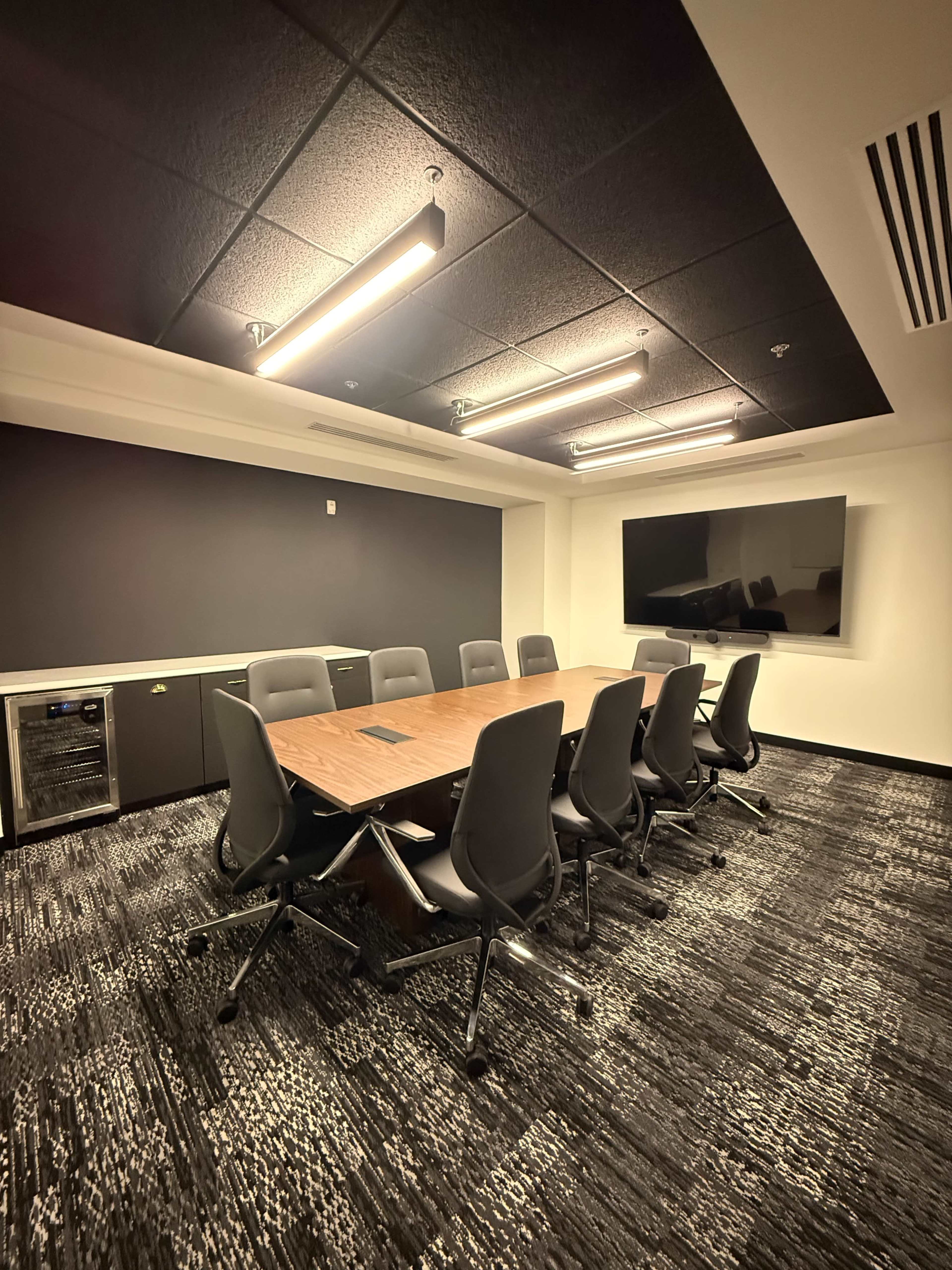 A modern conference room features a large wooden table surrounded by ergonomic chairs, with a flat-screen TV mounted on the wall and overhead lighting.