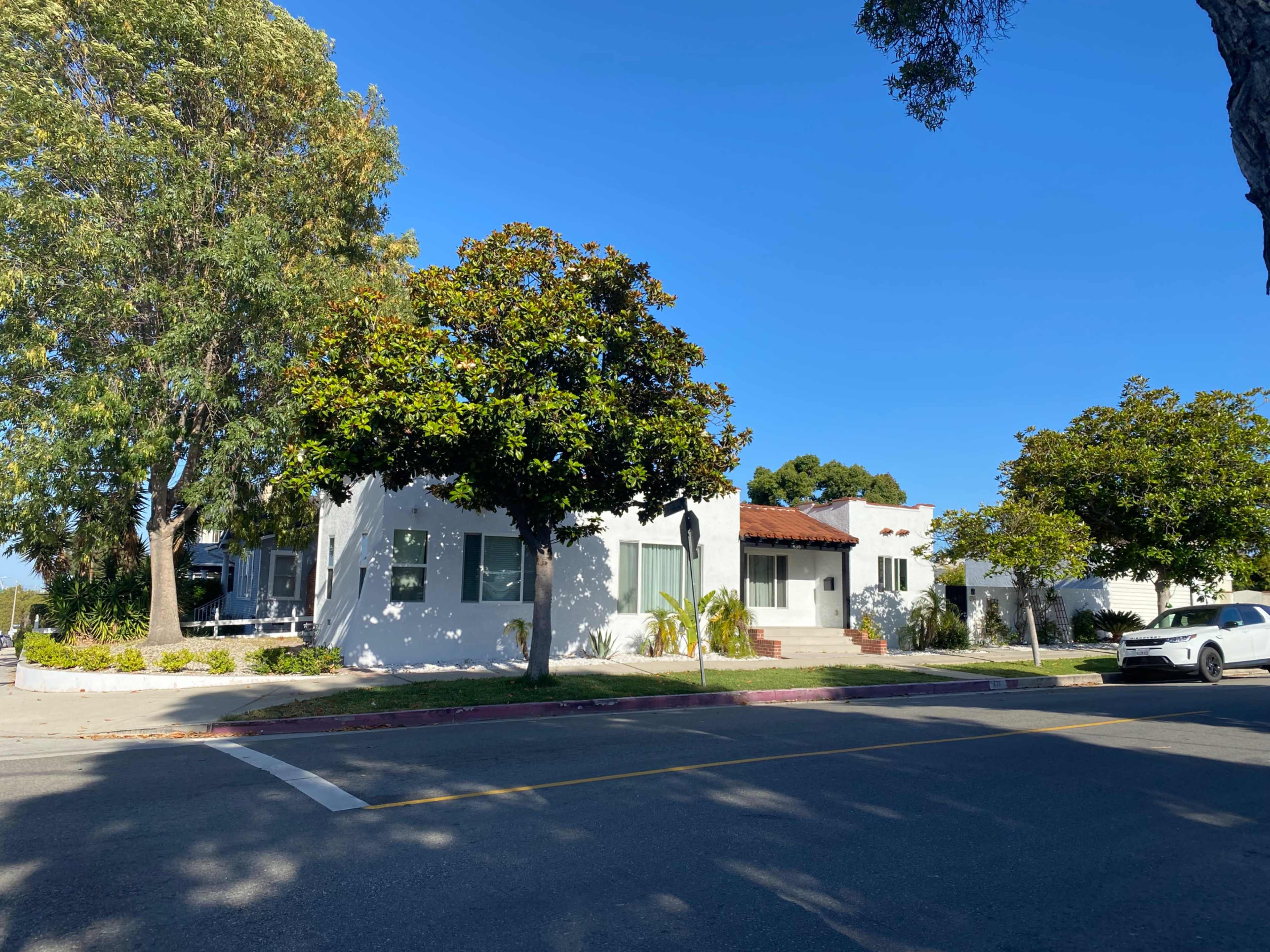 A white single-story house with a red tile roof and palm trees is located at the corner of a sunny street.