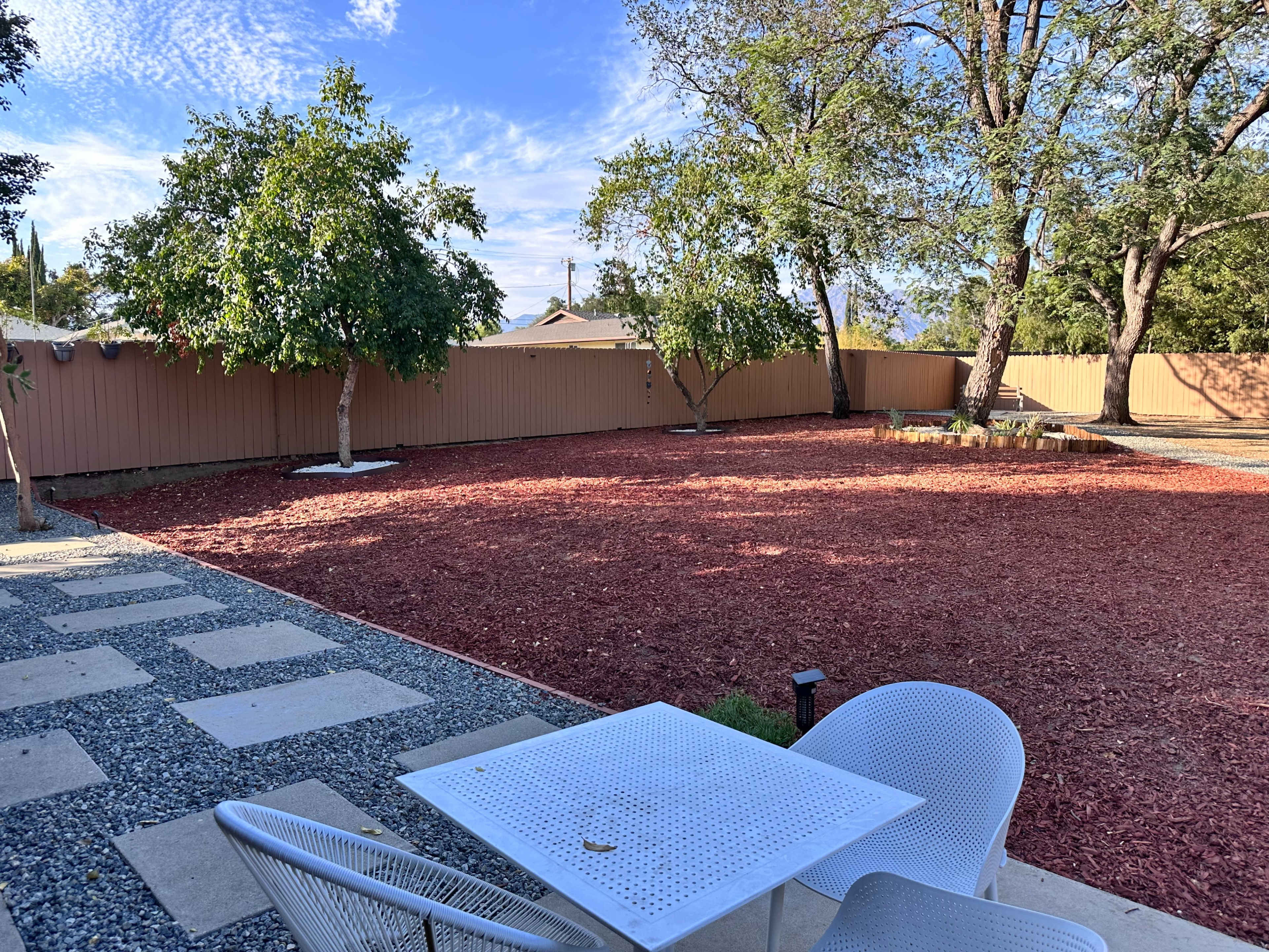 The image shows a landscaped backyard with red wood chips covering the ground, two trees, and a small patio area with a white metal table and chairs.