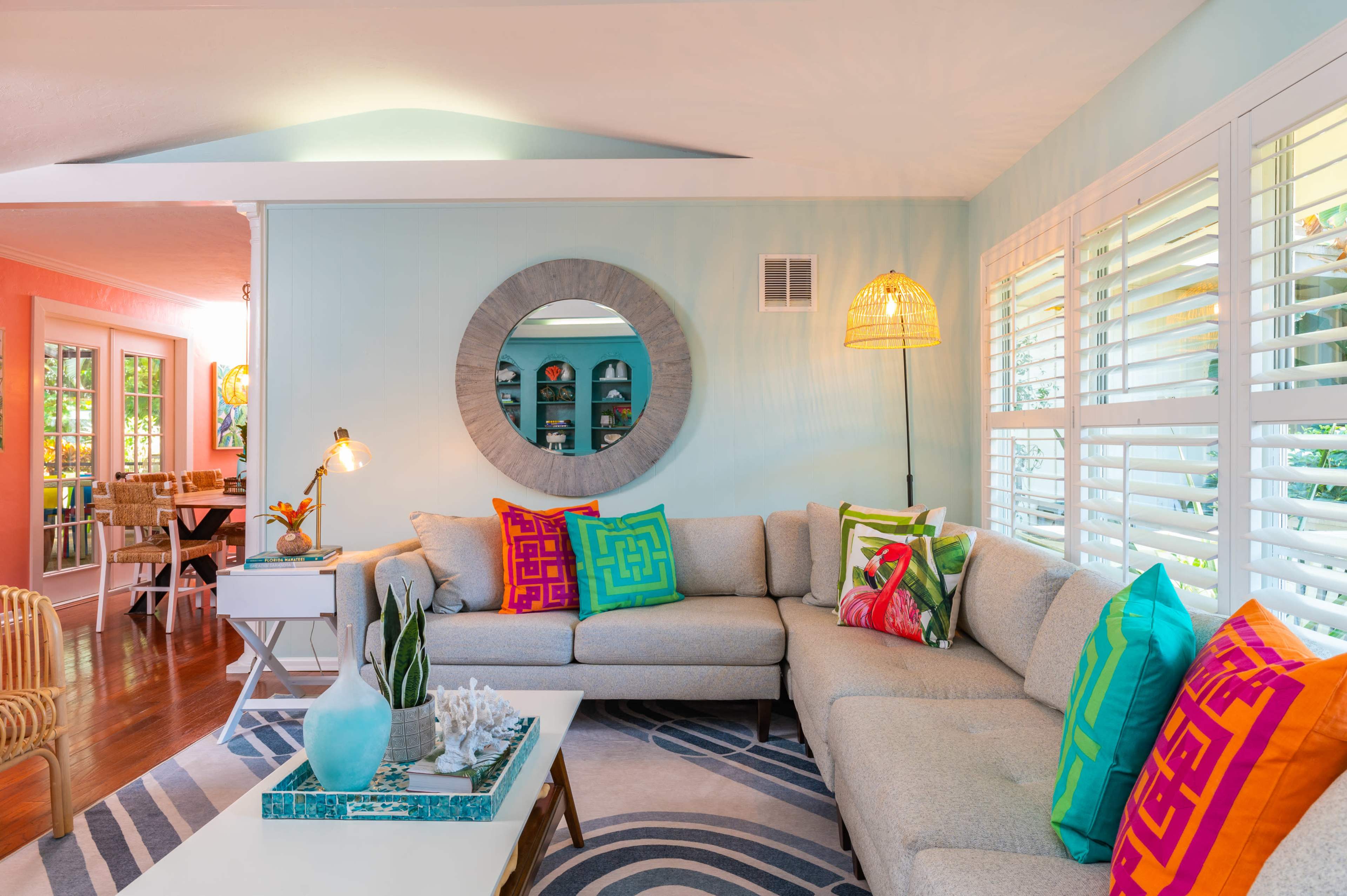 A light-colored living room features a sectional sofa adorned with colorful pillows, a round mirror on the wall, and a coffee table with decorative items.