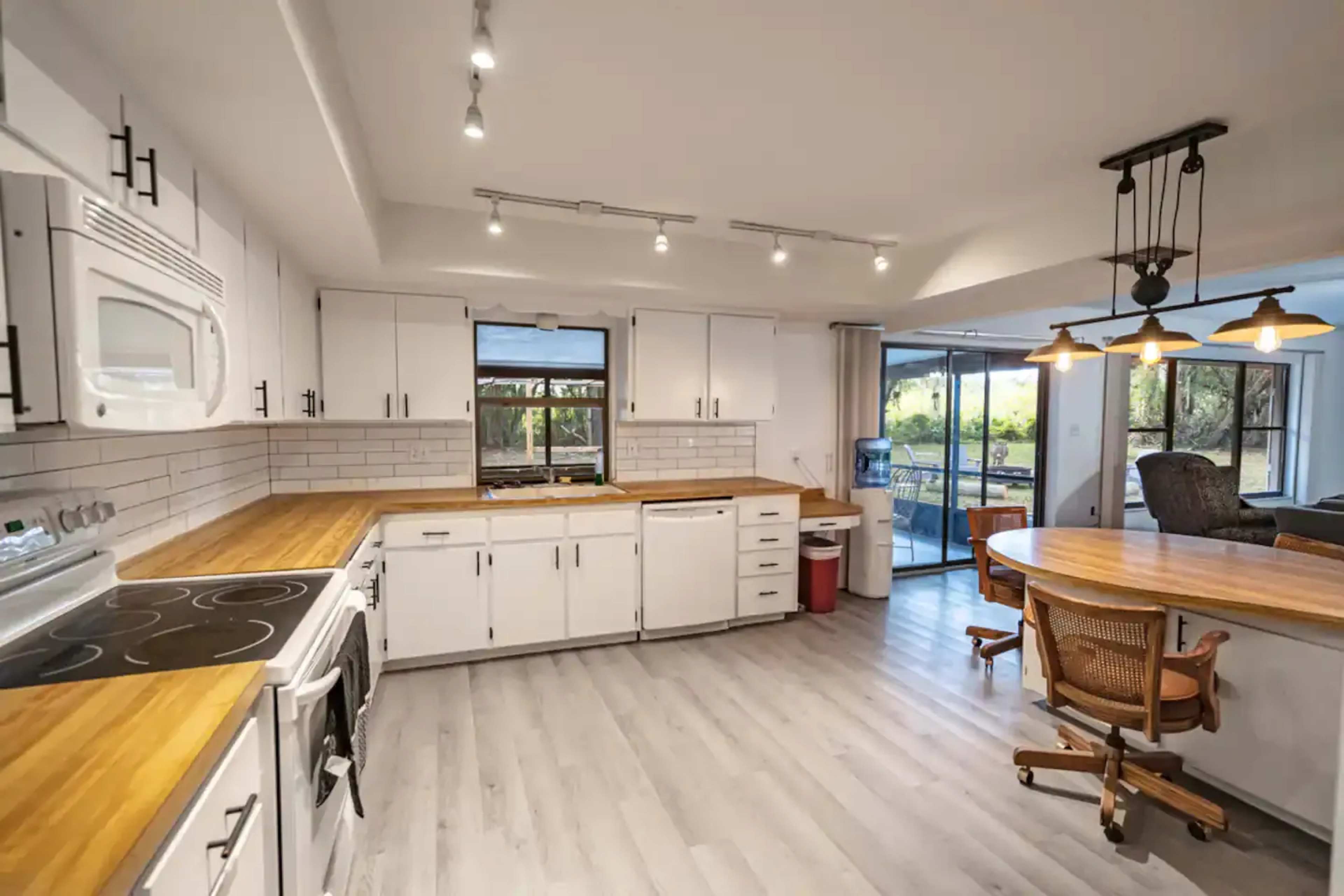 The image shows a spacious kitchen with white cabinets, wooden countertops, modern appliances, and a dining area featuring a round table.