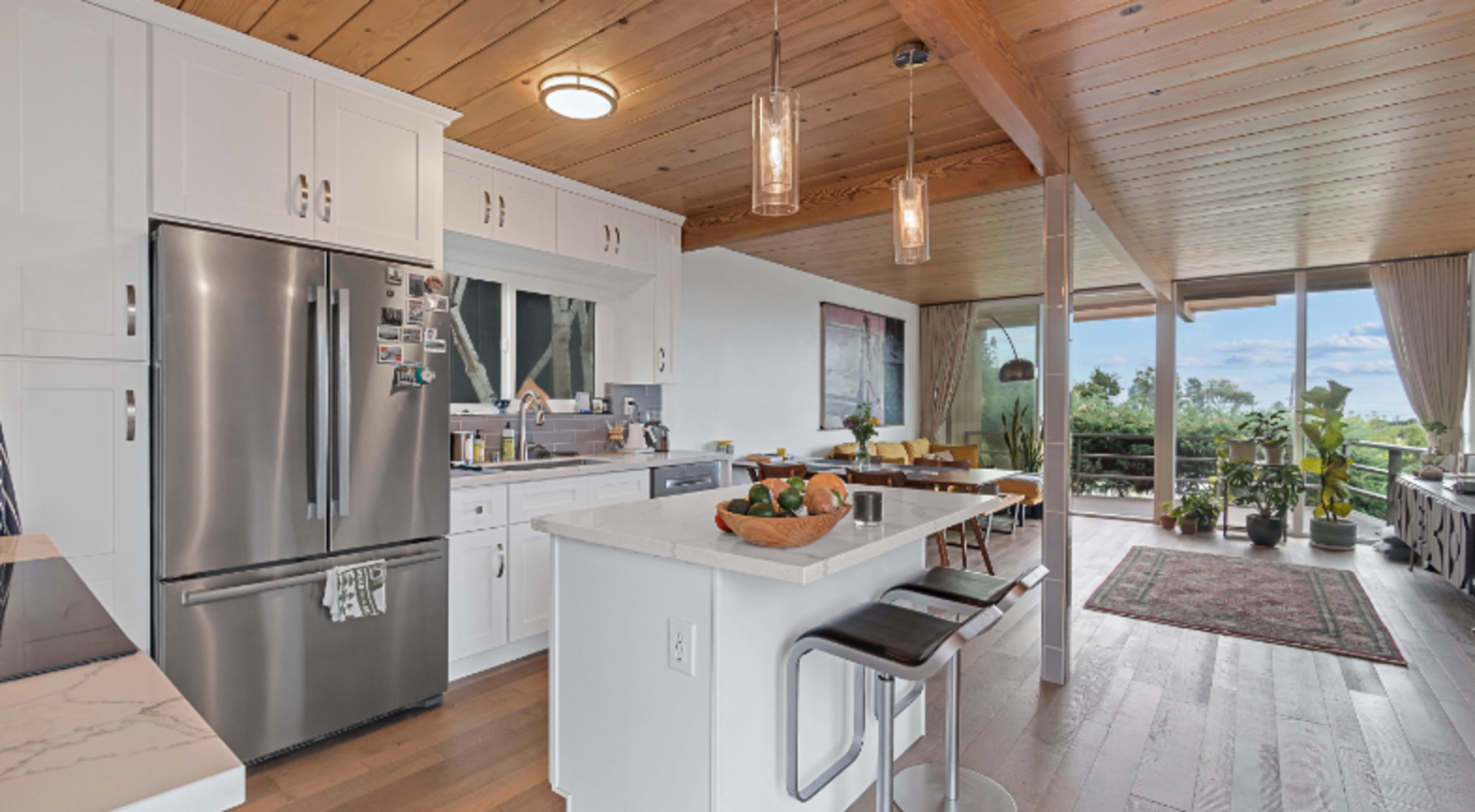 A modern kitchen features stainless steel appliances, white cabinetry, and an island with seating, open to a dining area with large windows and plants.