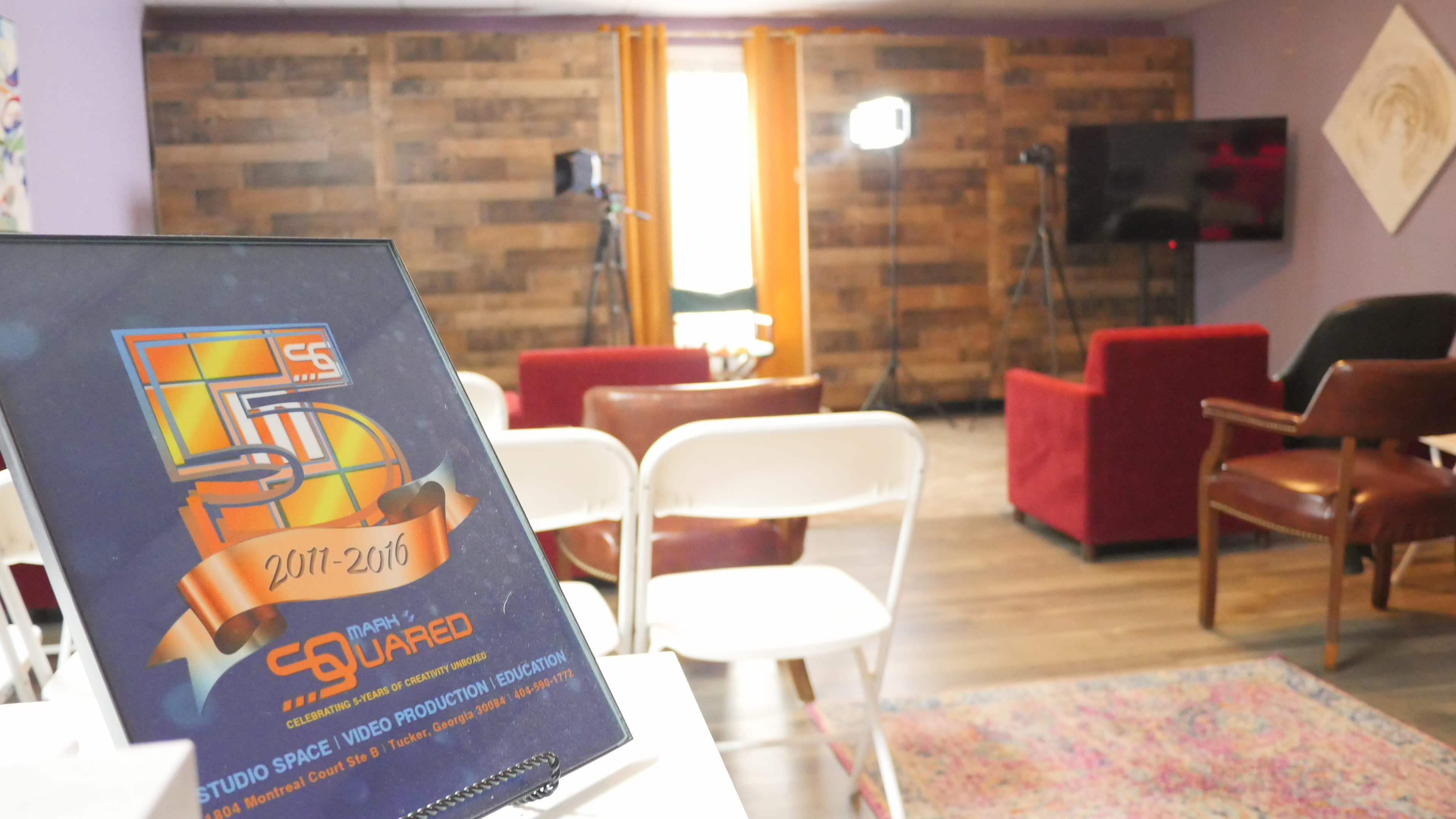 A cozy studio space set up for video production, featuring a decorative plaque in the foreground, chairs arranged for an audience, and lighting equipment positioned in the background.