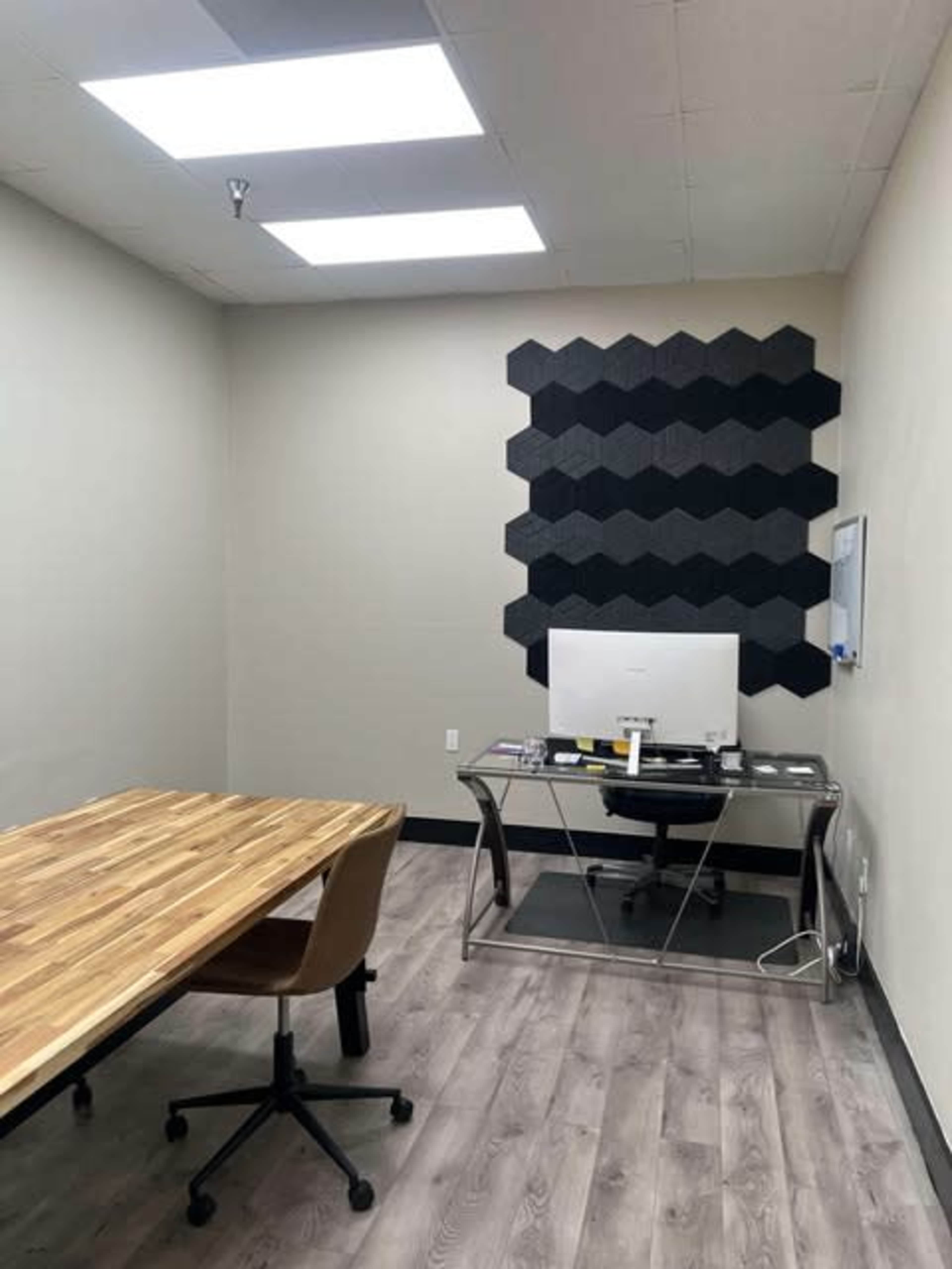 The image shows a minimalist office space with a wooden conference table, an office chair, and a glass desk that holds a computer, against a wall featuring a hexagonal black panel design.