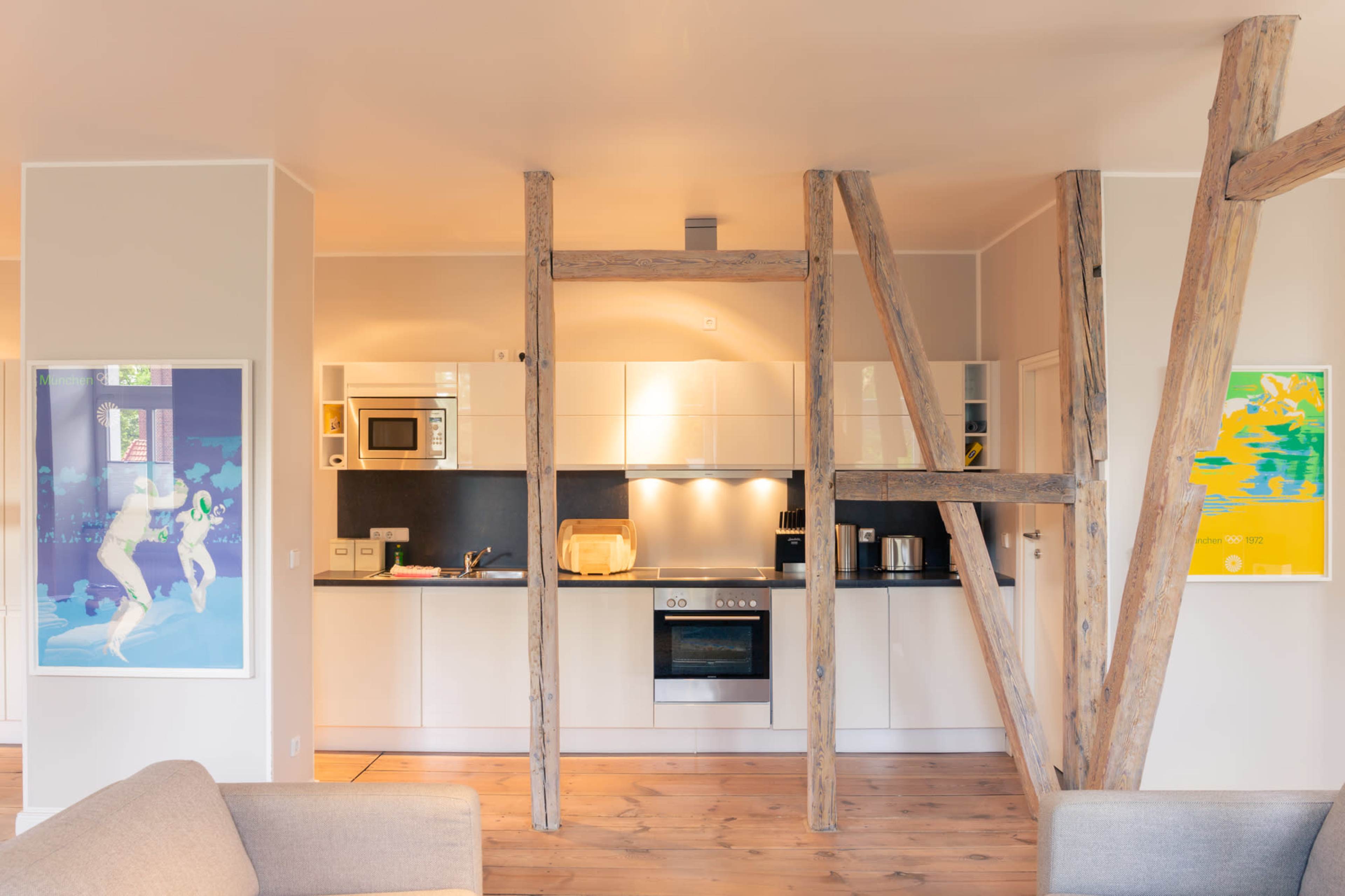 A modern kitchen with wooden support beams, featuring white cabinets, a stove, a microwave, and colorful artwork on the walls.