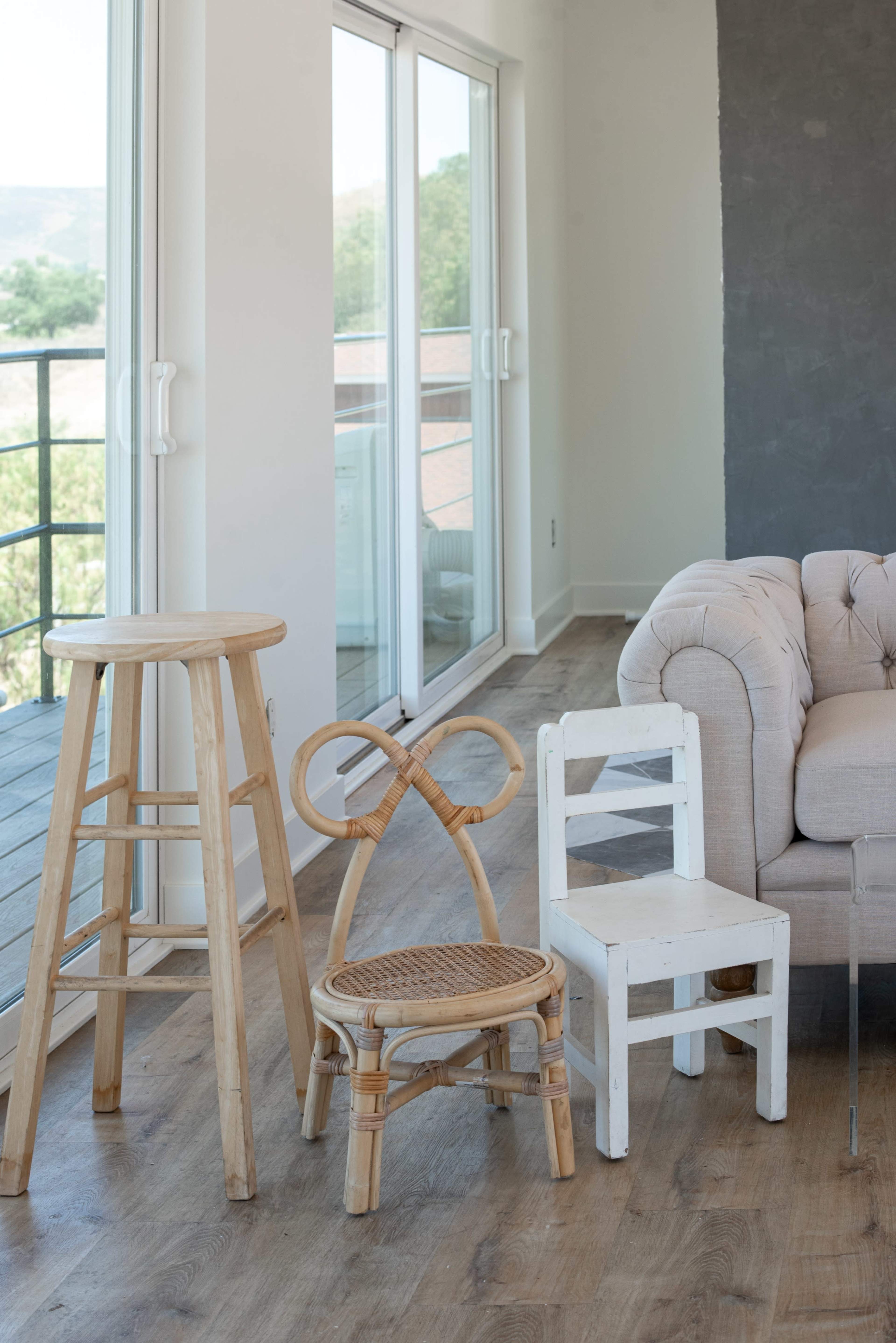 The image shows a modern interior with a wooden floor, featuring a beige sofa and three distinct chairs: a tall wooden bar stool, a woven chair with a decorative backrest, and a simple white wooden chair.