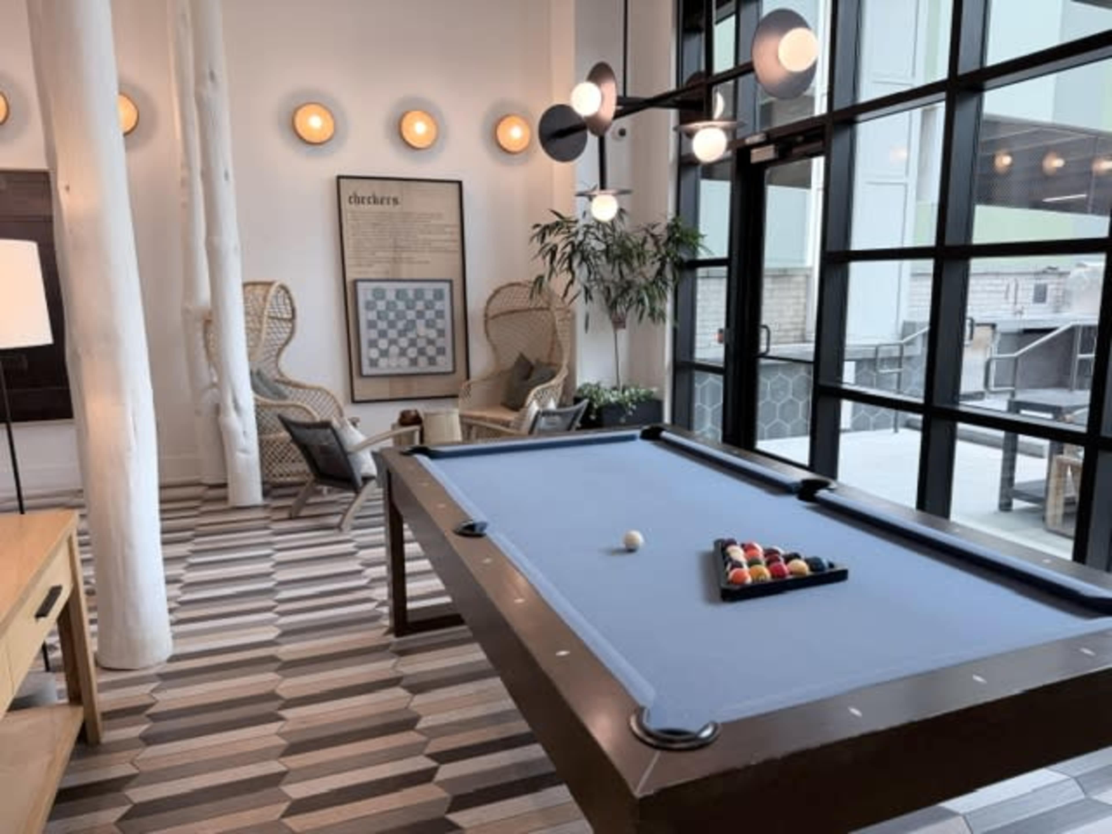 The image shows a recreational room featuring a pool table with balls arranged on it, surrounded by chairs and a large window letting in natural light.