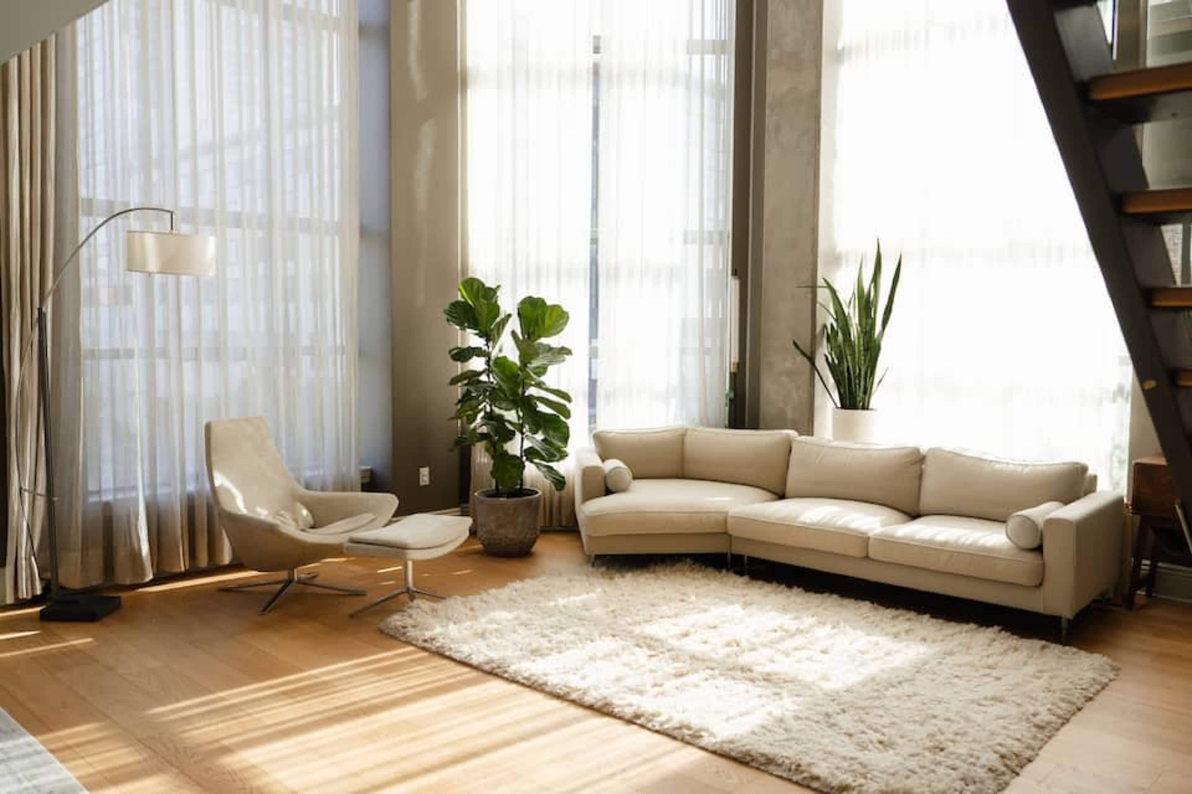 A modern living room features a light-colored sofa, an armchair, potted plants, and large windows with sheer curtains allowing natural light to fill the space.