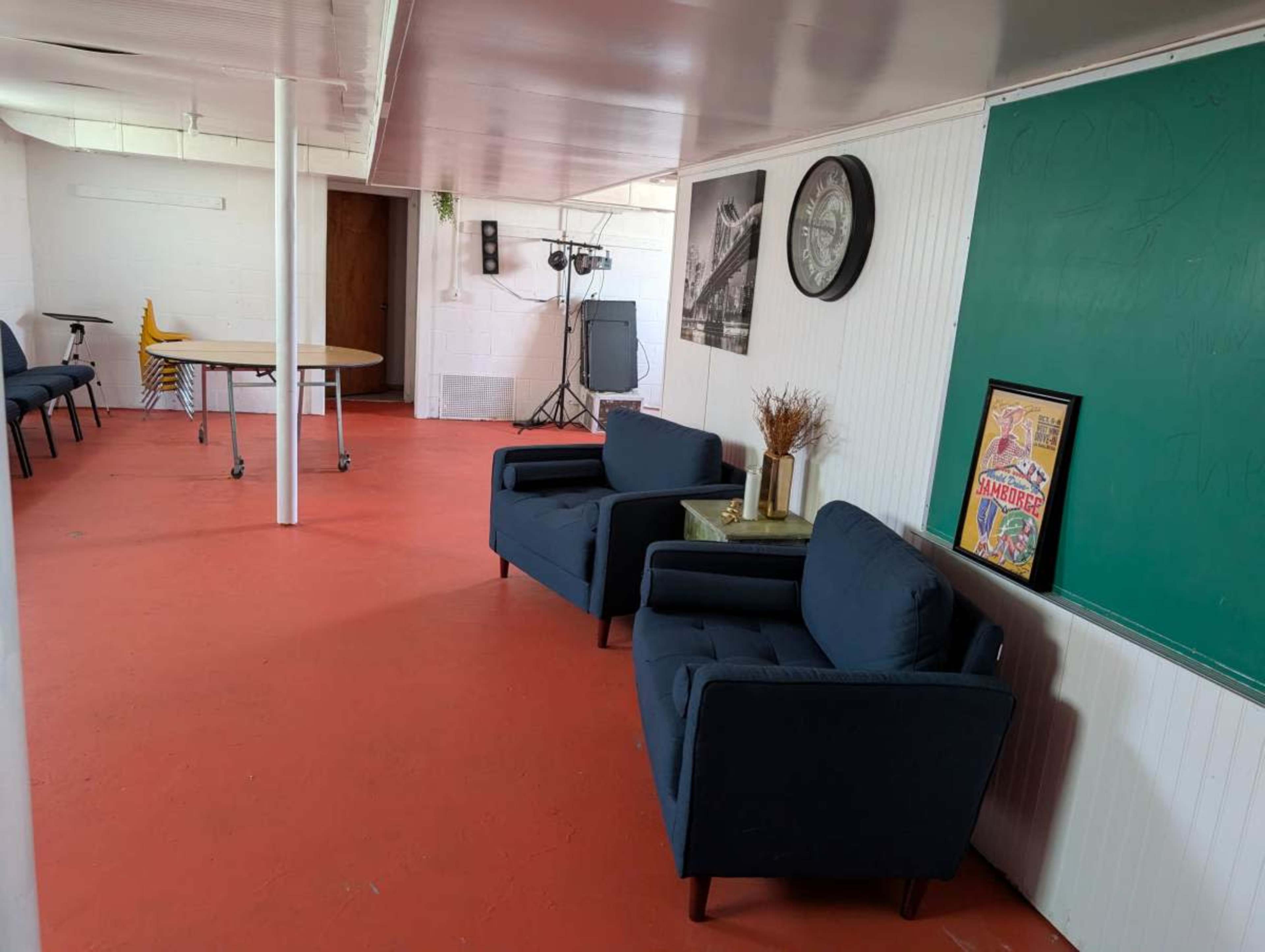 The image shows a room with red flooring, two blue chairs, a small table, a chalkboard, and a wall clock, along with some lighting equipment in the background.
