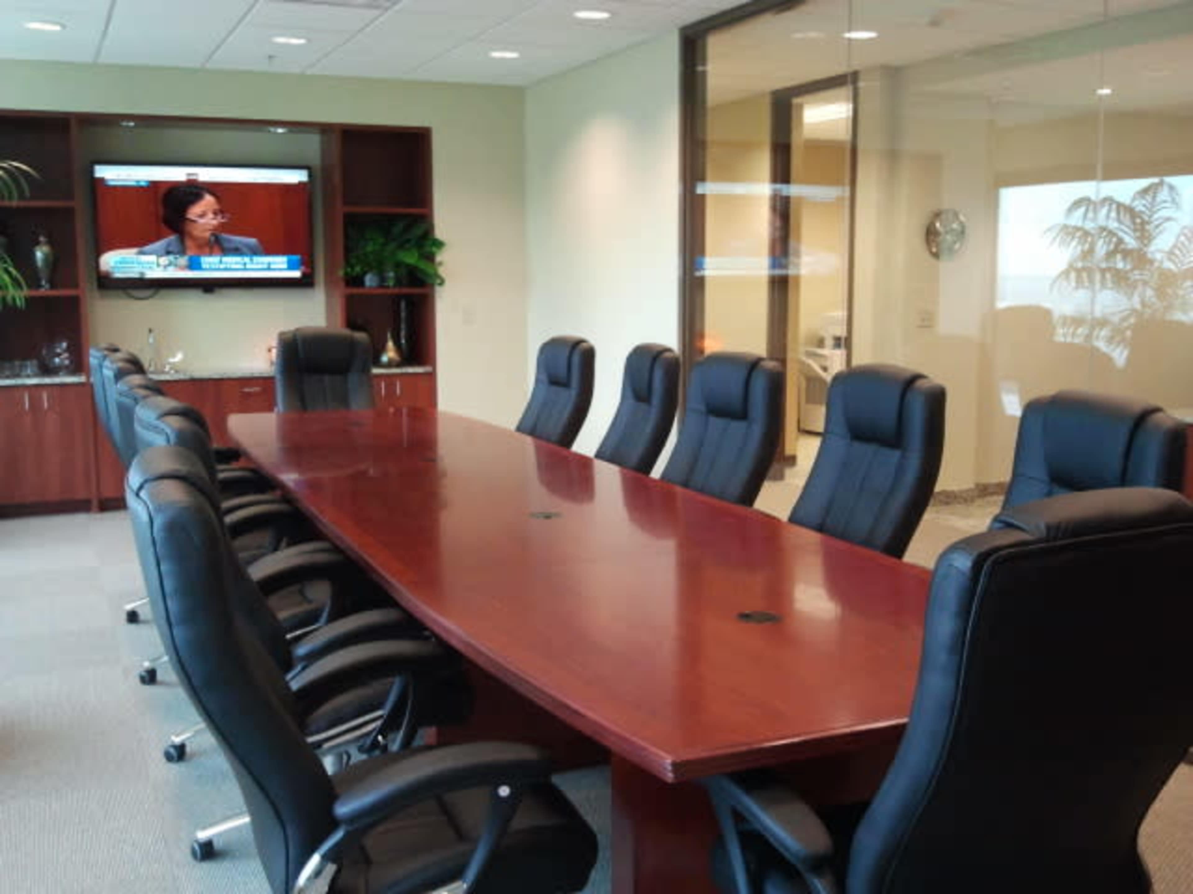 The image shows a conference room with a large wooden table surrounded by eight black chairs, and a television displaying a news segment mounted on the wall.