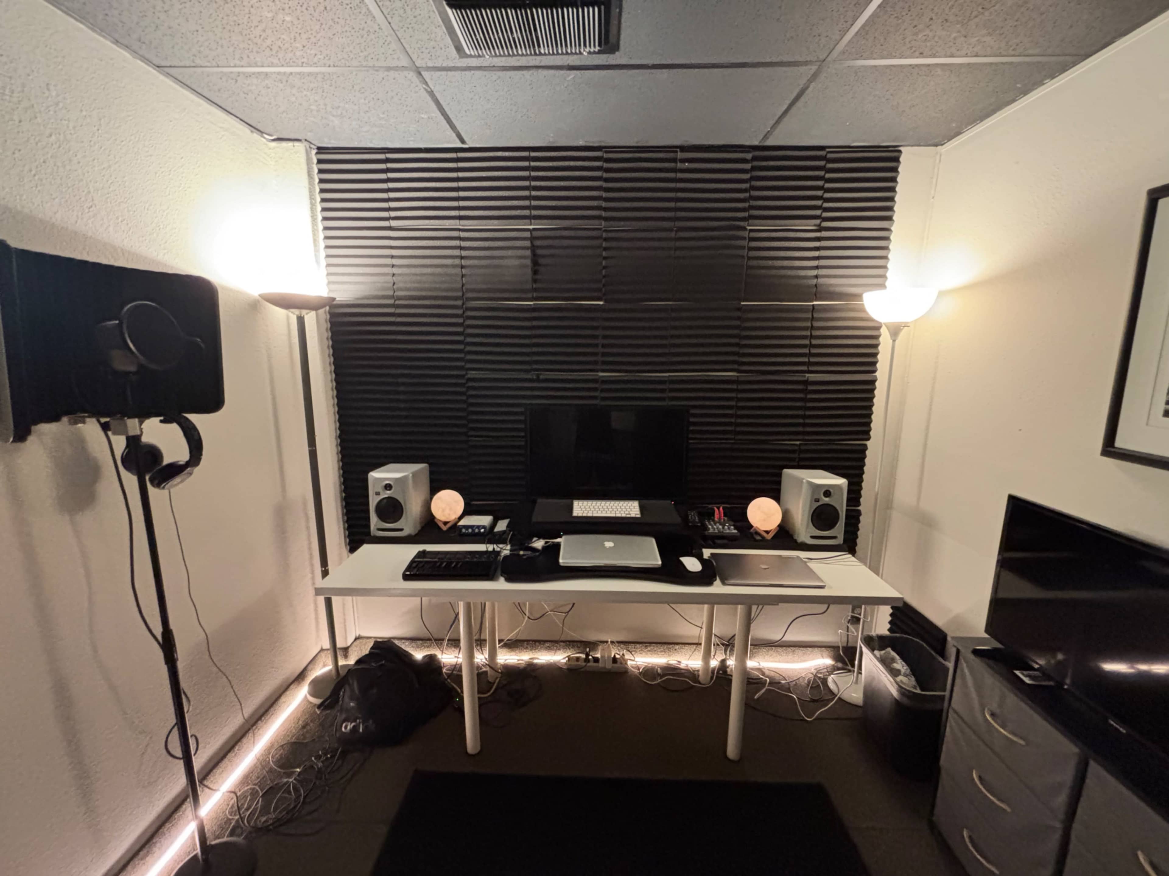 The image shows a music production studio with a computer setup, speakers, a microphone stand, and soft lighting against black acoustic paneling.