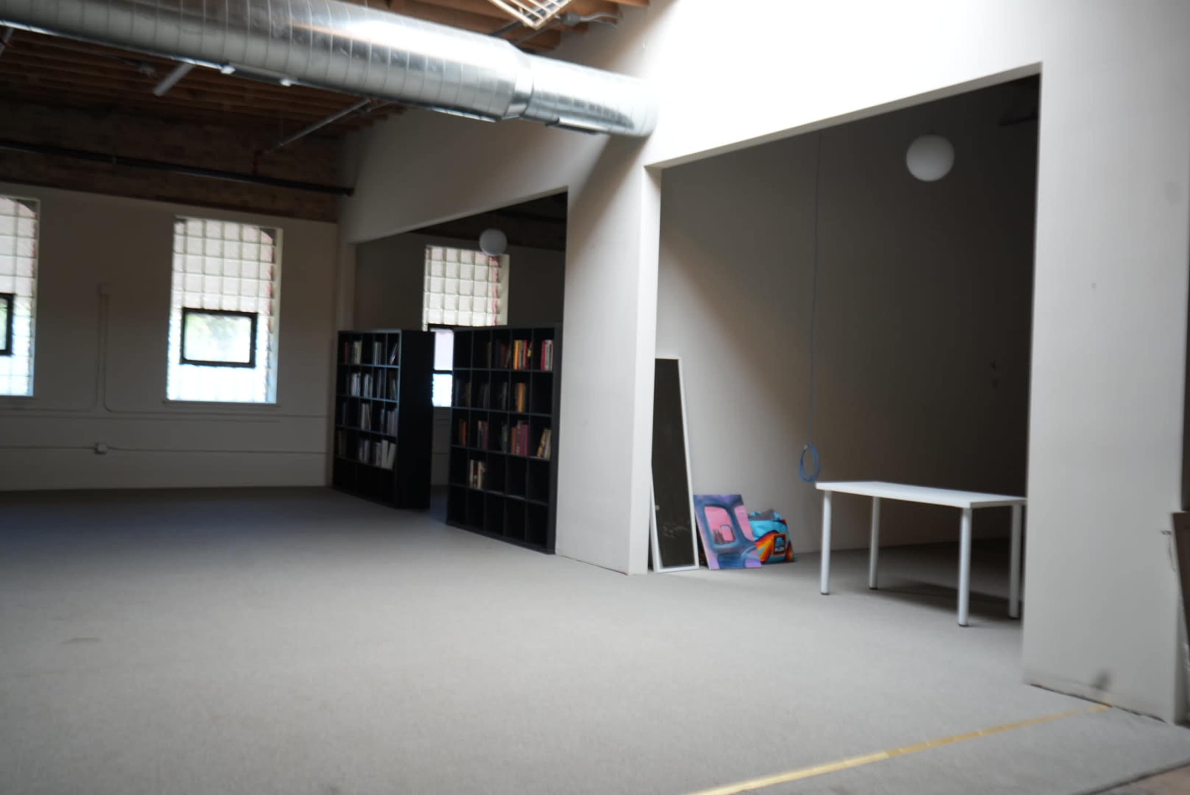 The image shows a spacious, industrial-style room with a carpeted floor, featuring two openings leading to adjacent areas, a bookshelf filled with books, a mirror, and a white table.