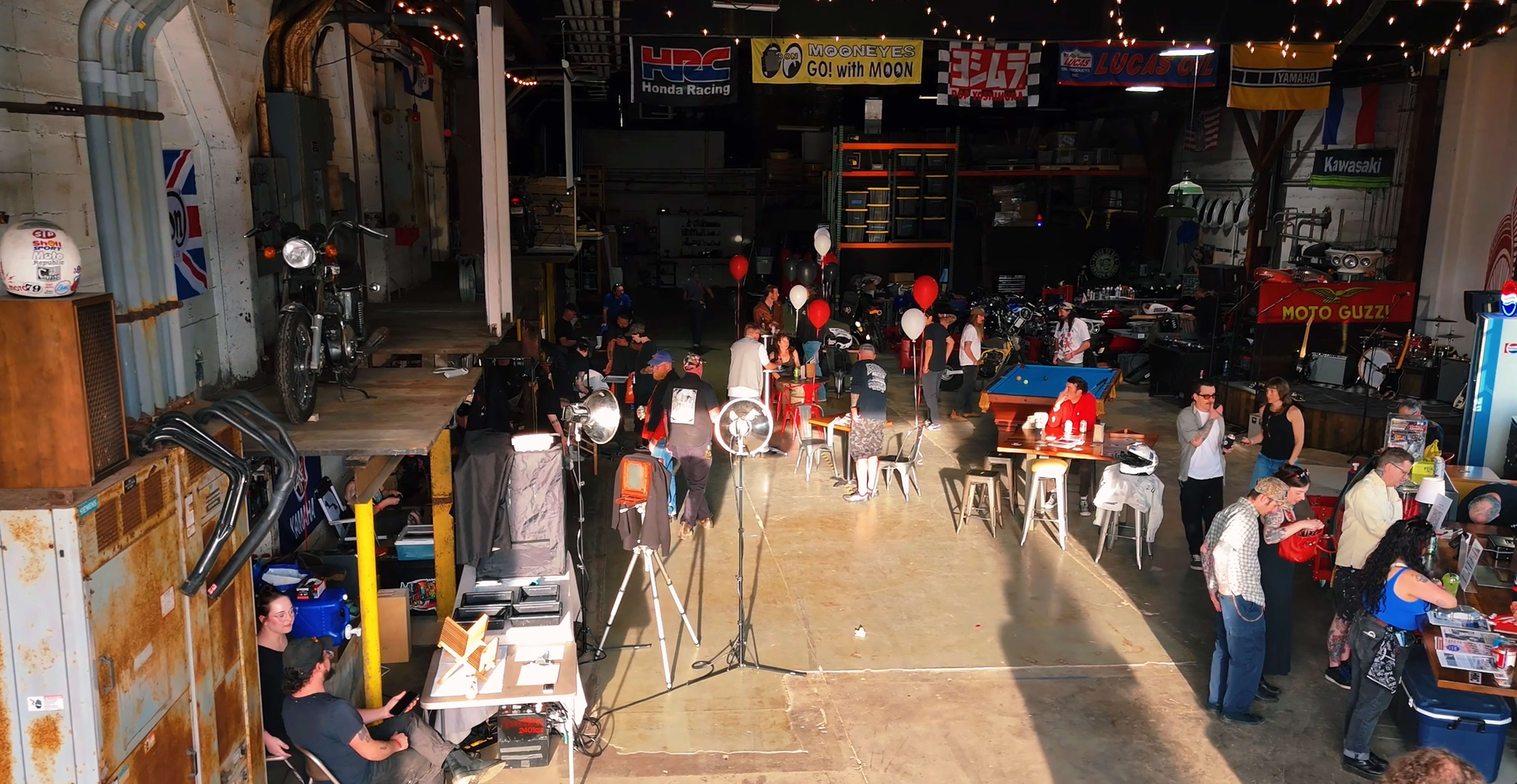 The image shows a bustling workshop or event space filled with people, tables, and decorations, alongside various motorcycles and equipment.