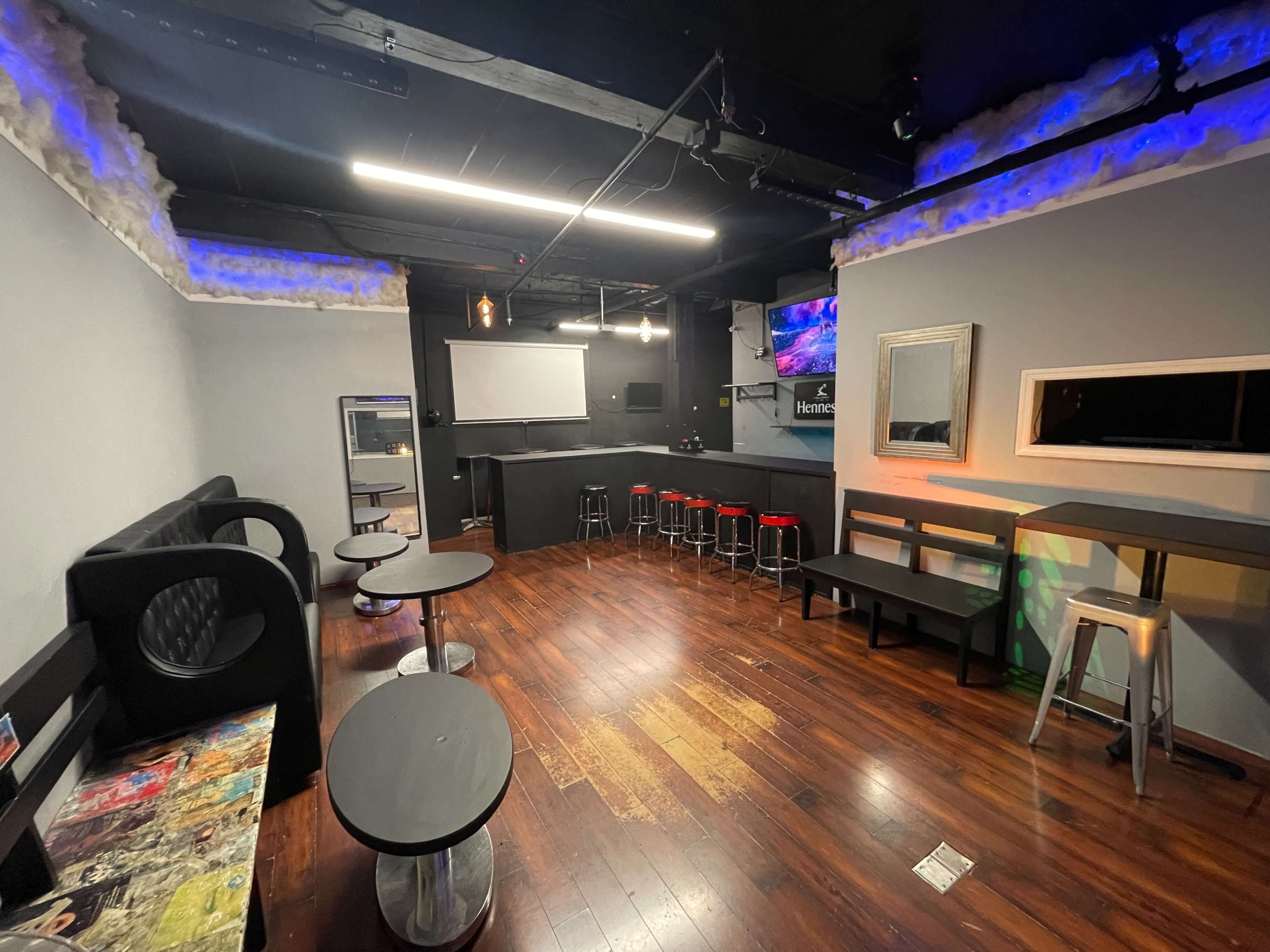 A dimly lit lounge area featuring black seating, small tables, and a bar with red stools against a backdrop of gray walls.