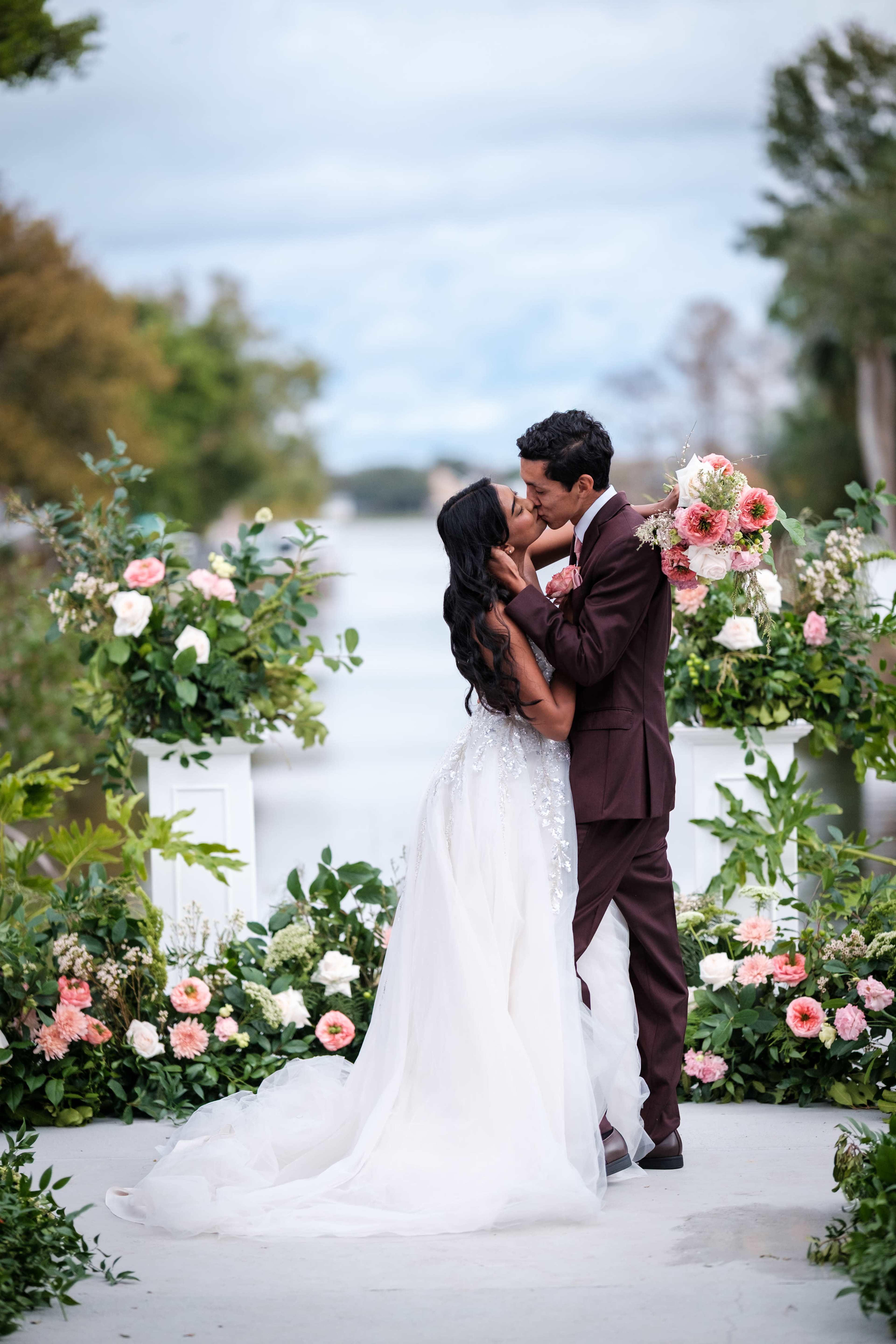 A bride and groom kiss on a pathway adorned with floral arrangements by a waterway.