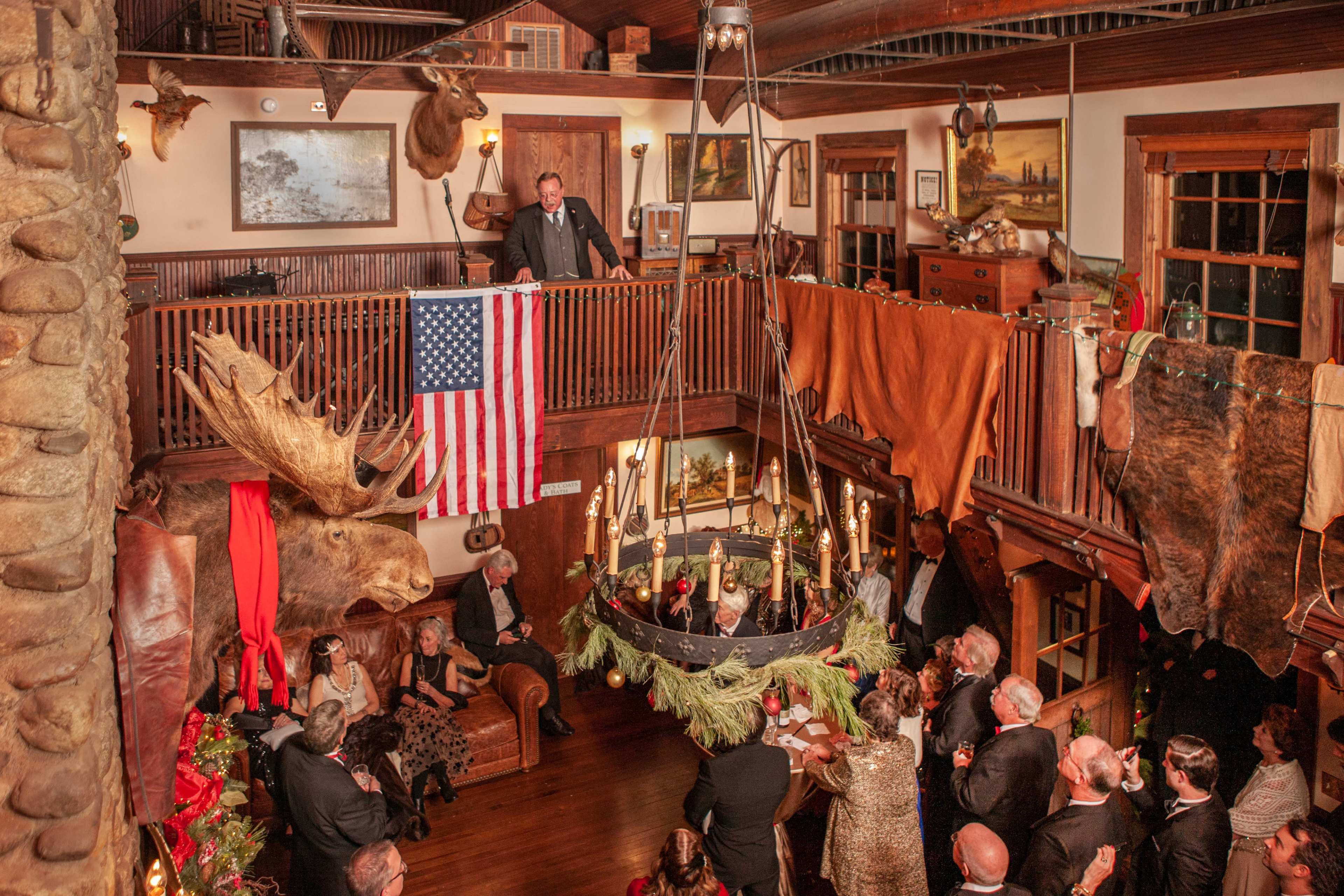 A large gathering takes place in a rustic lodge, featuring taxidermy mounts, an American flag hanging from the balcony, and guests dressed in formal attire.