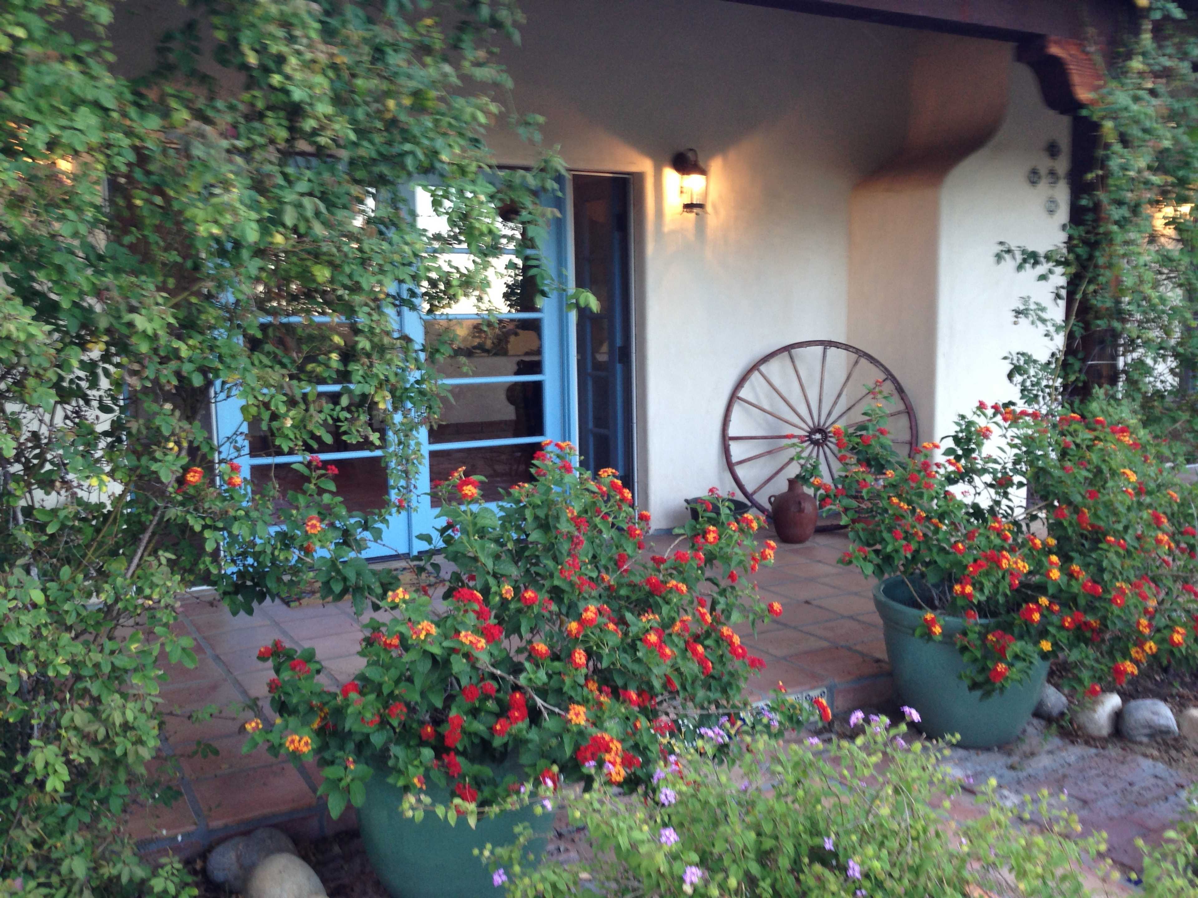 The entrance features a blue door surrounded by vibrant flowers in large pots, with a wooden wagon wheel mounted on the wall nearby.