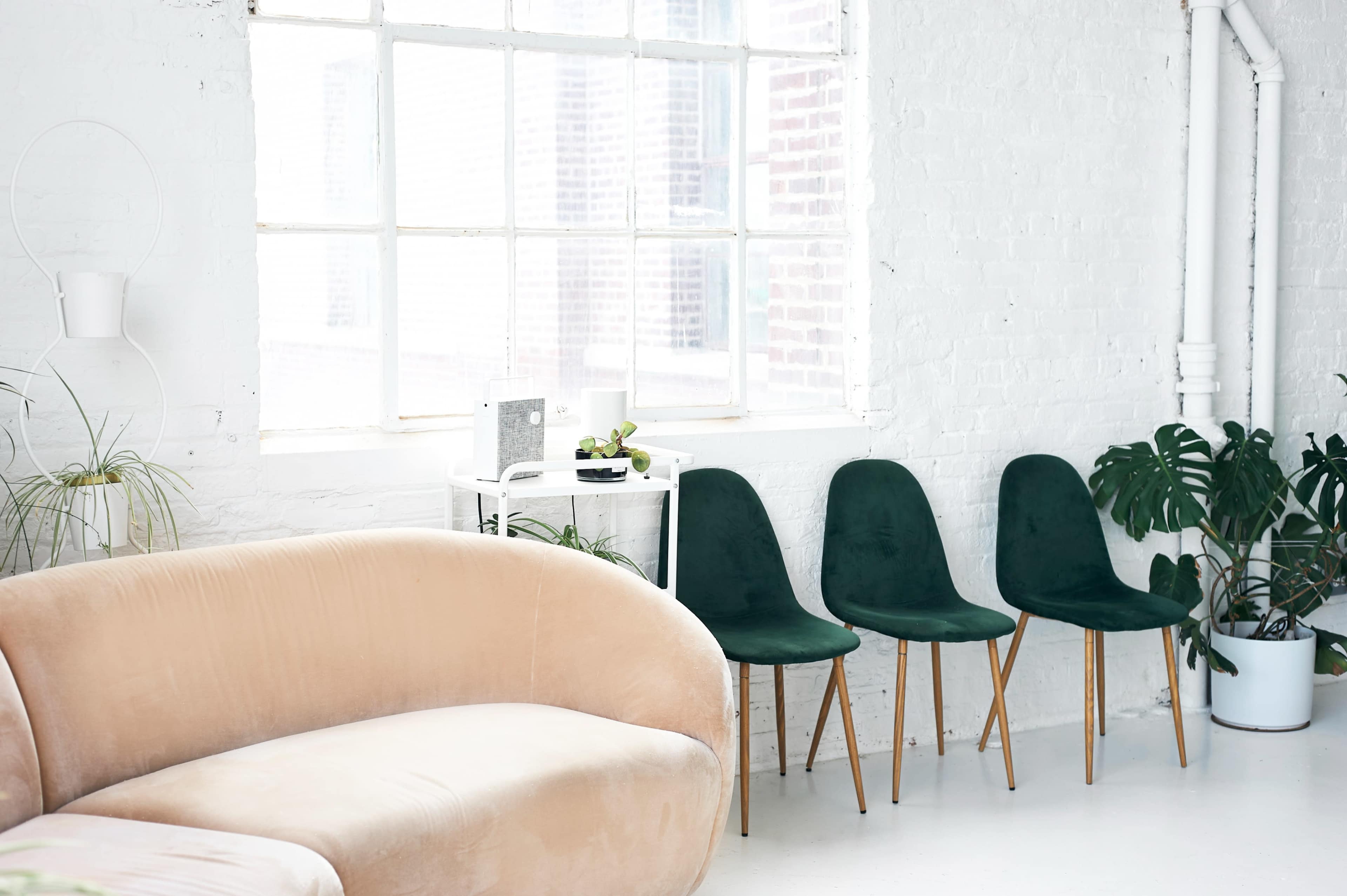 The scene features a modern, bright room with a light-colored curved sofa and several green chairs positioned against a wall with large windows.