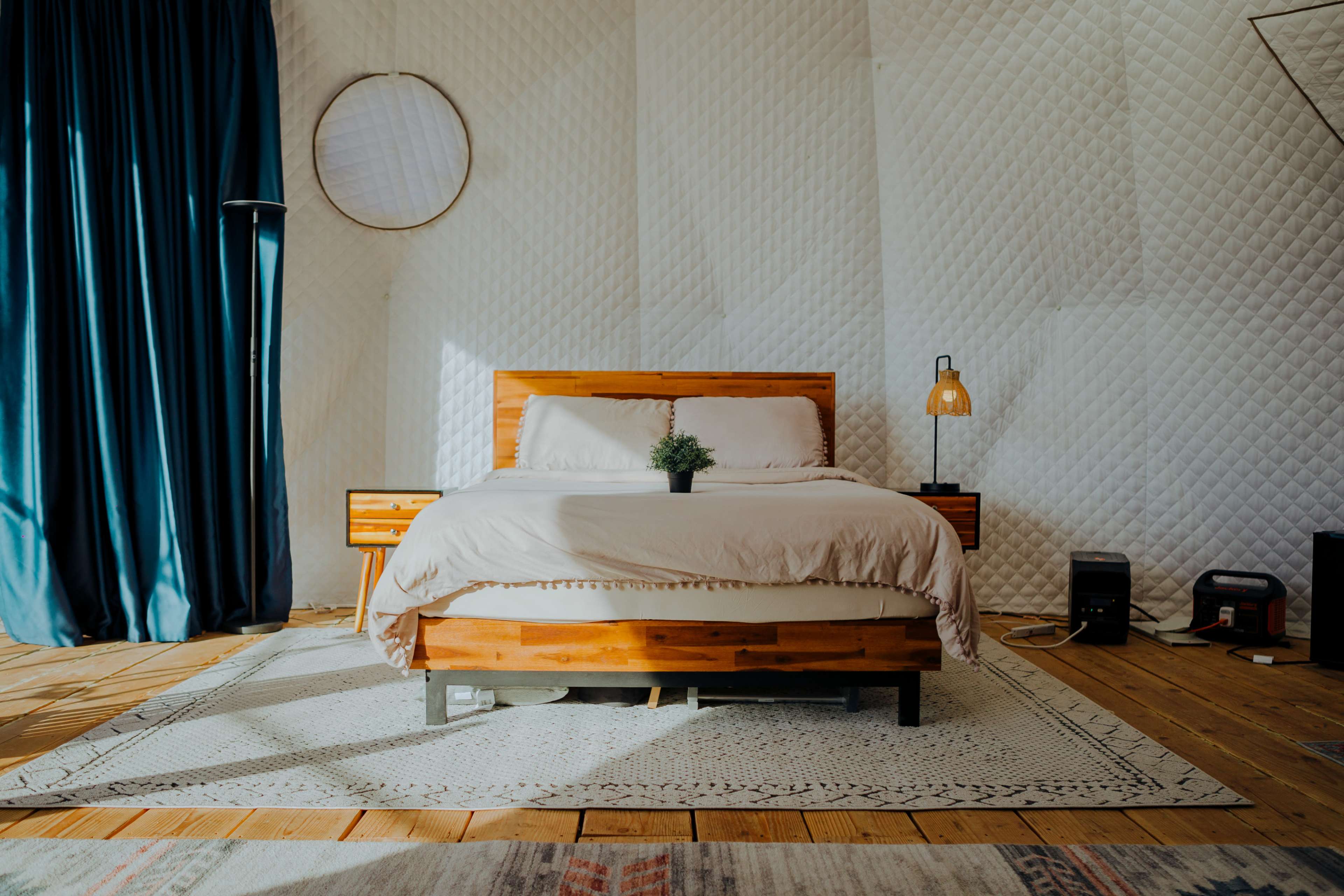 The image shows a neatly arranged bedroom featuring a wooden bed with a light-colored blanket, a bedside table, and a decorative plant, all set against a backdrop of textured white walls and dark curtains.