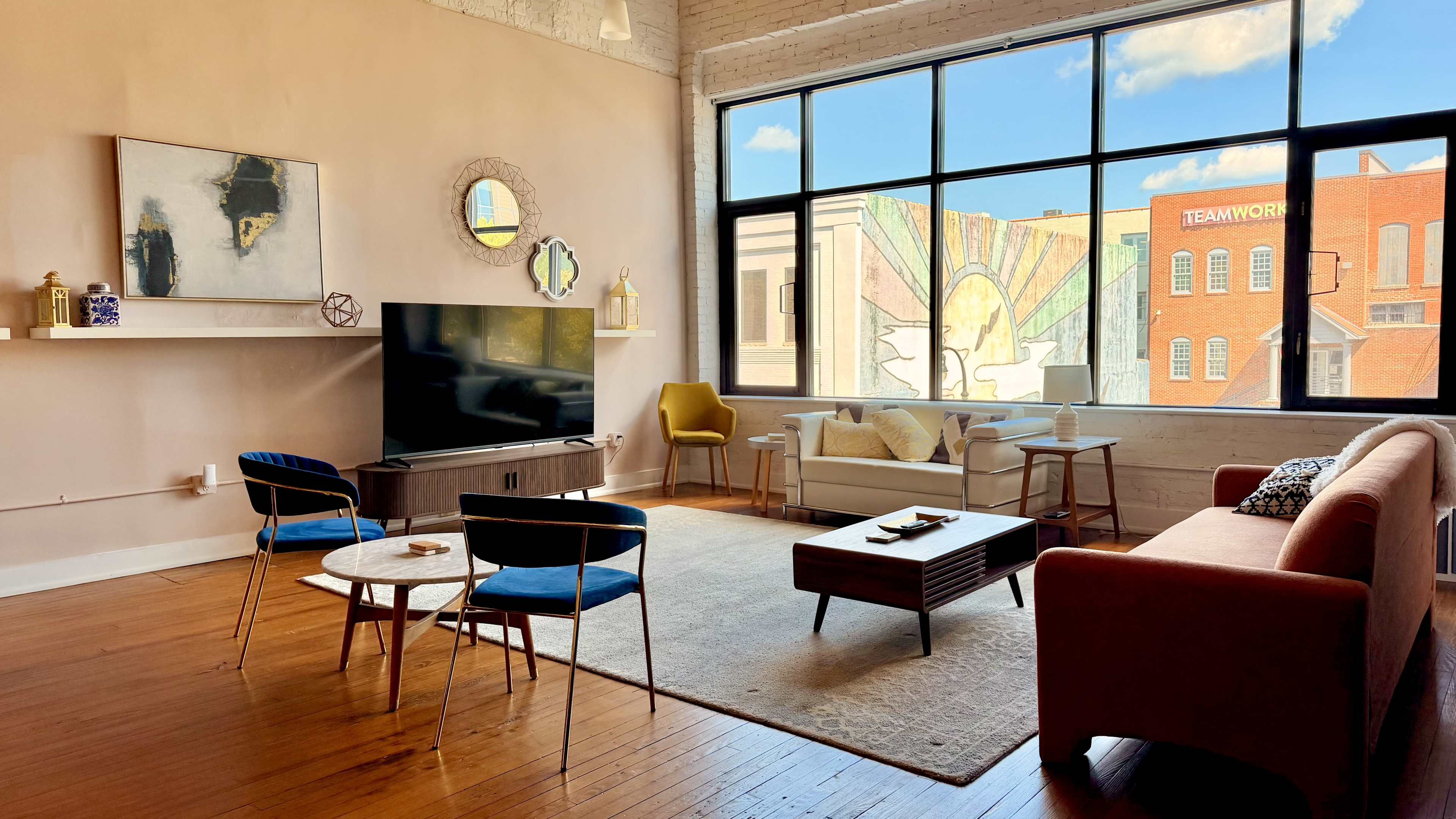 The scene depicts a modern living room with large windows, furnished with a TV, a sofa, armchairs, and a coffee table, complemented by decorative wall art.