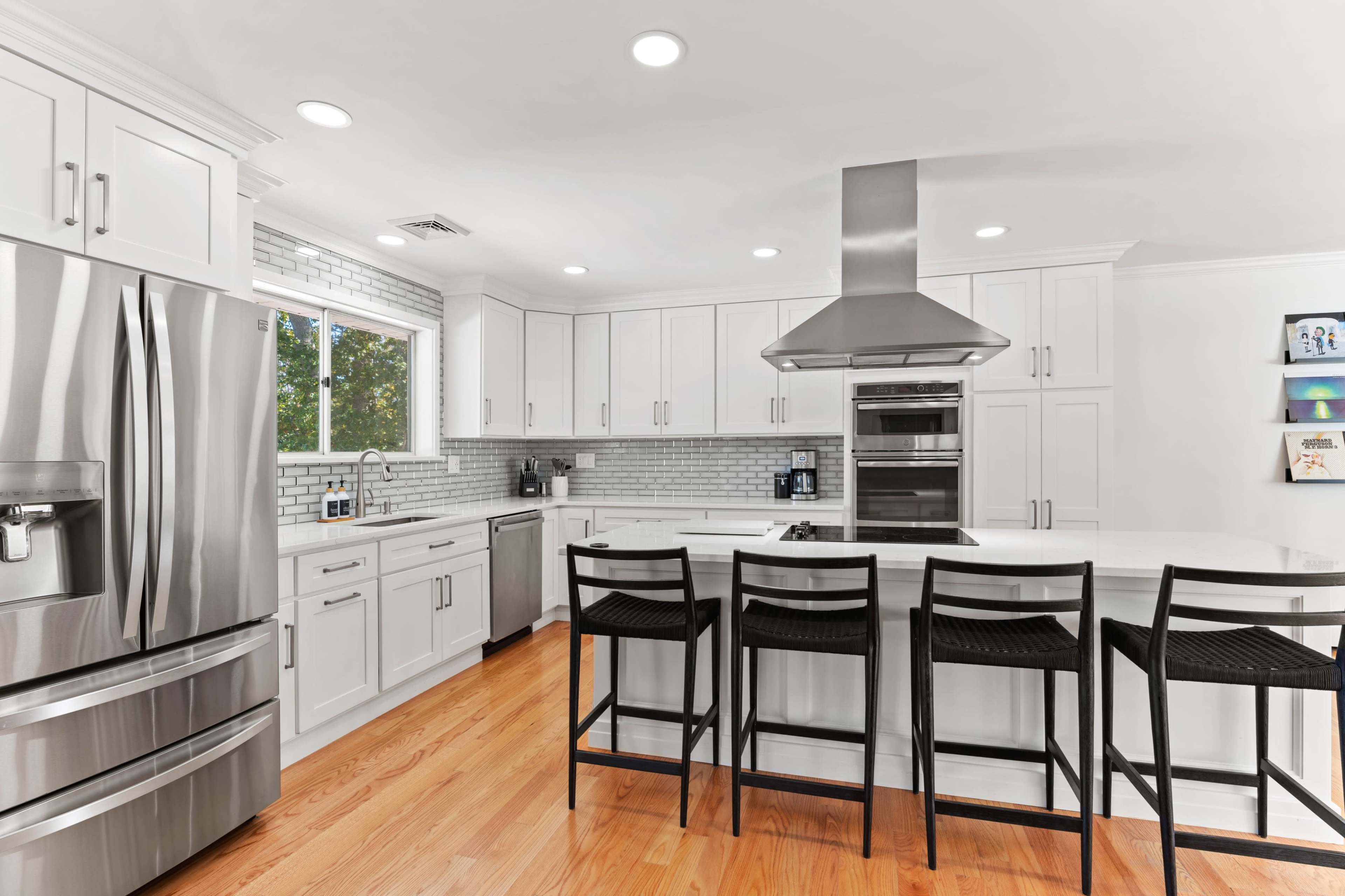 The image shows a modern kitchen with white cabinetry, a large island with bar stools, stainless steel appliances, and a backsplash of light gray tiles.