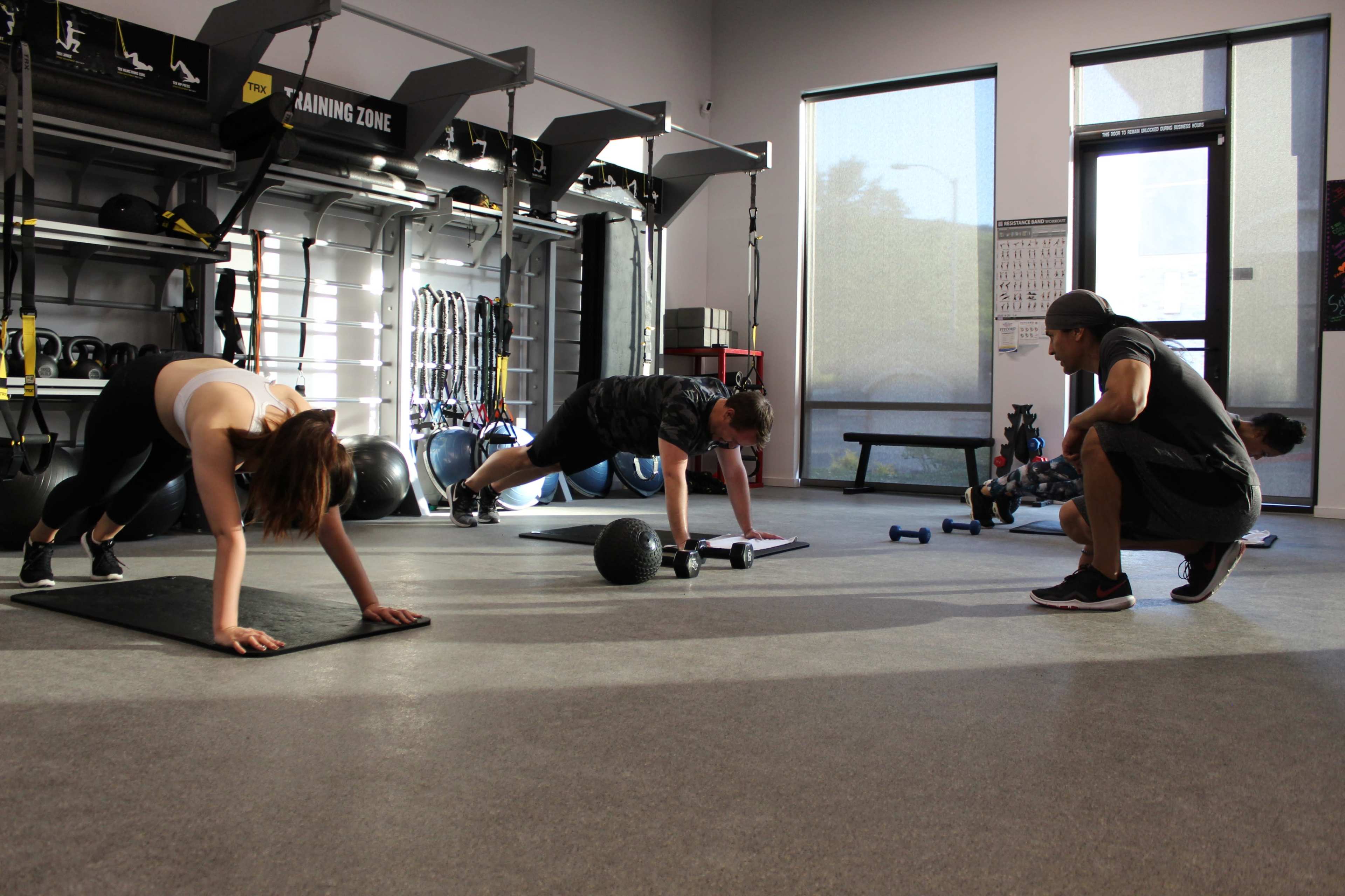 Two individuals perform exercises on mats in a gym while a trainer observes them.