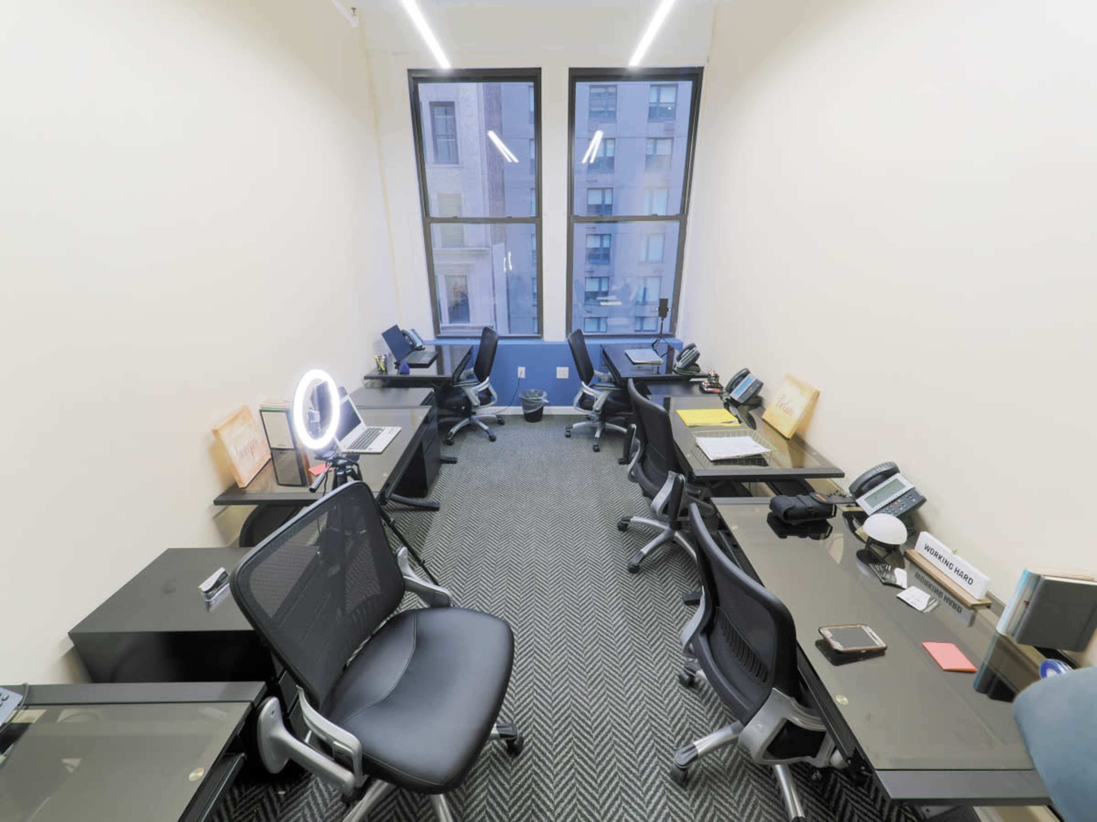 A modern office space features several desks equipped with computers, chairs, and phones, arranged in a well-lit, minimalist layout.