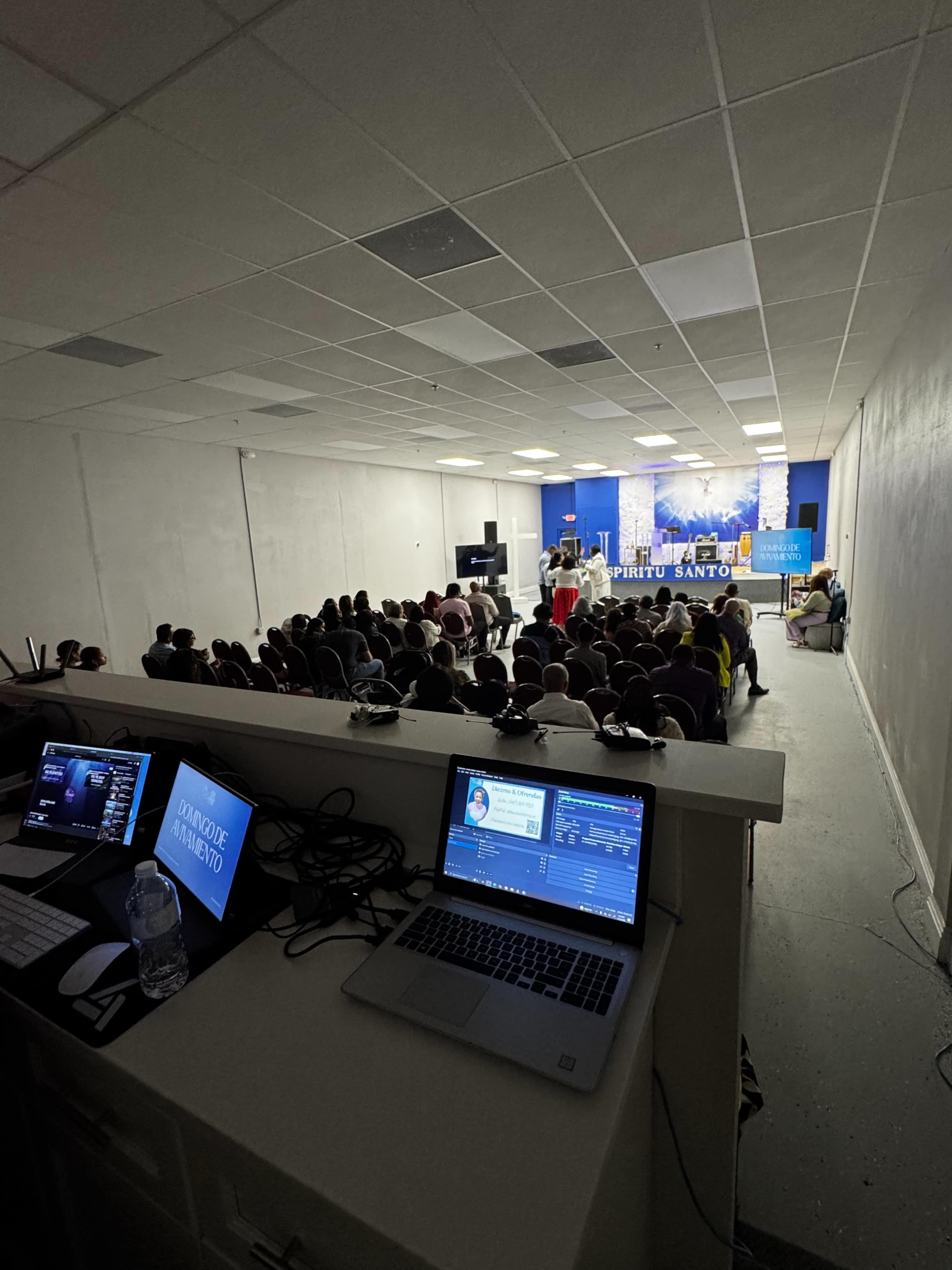 A group of people is seated in a church or meeting space, while individuals at the front conduct a presentation or service, with equipment visible in the foreground.