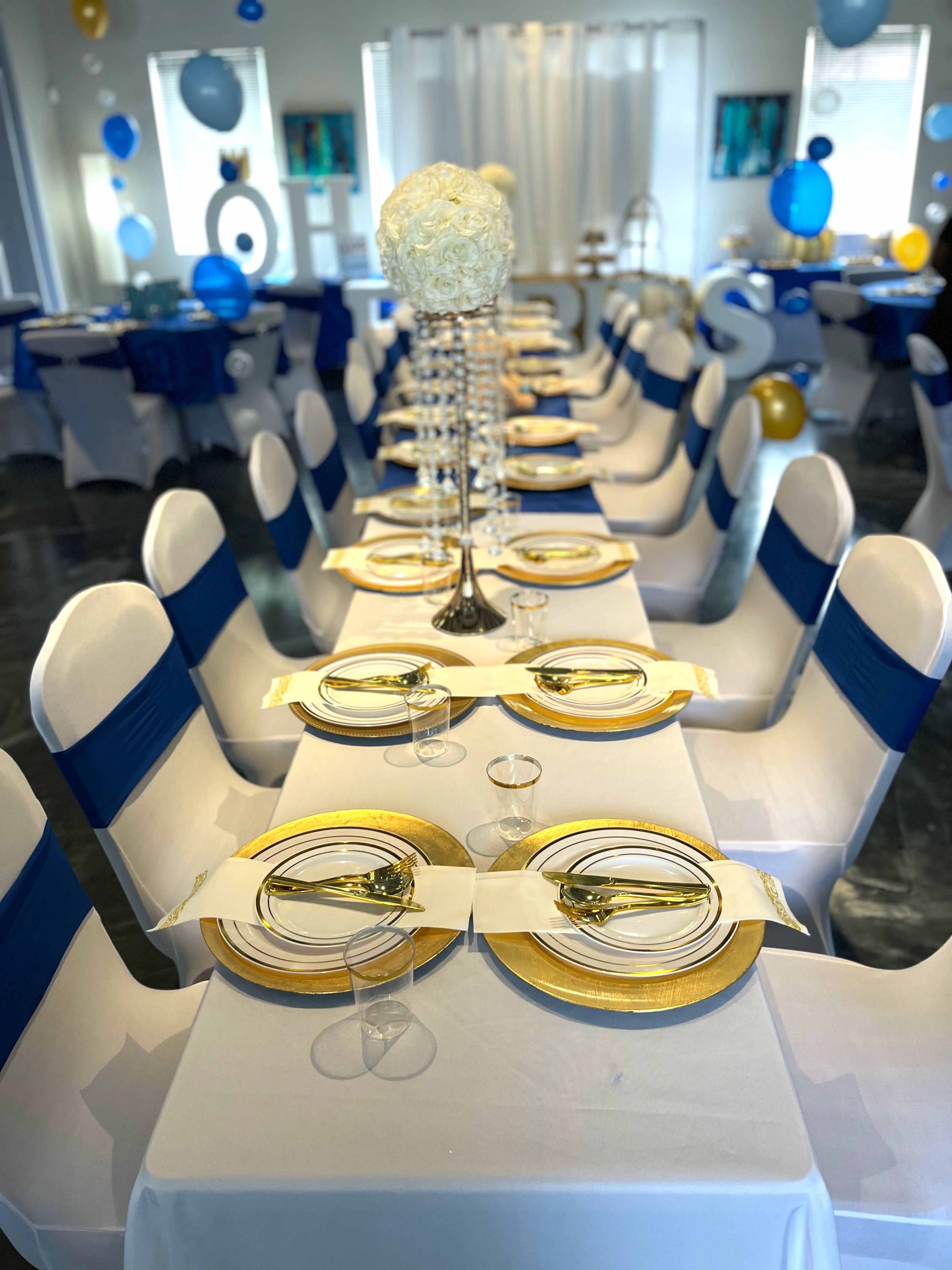 A banquet hall is set for an event with neatly arranged tables draped in white cloths and accented with blue bows, featuring gold rimmed plates and floral centerpieces.