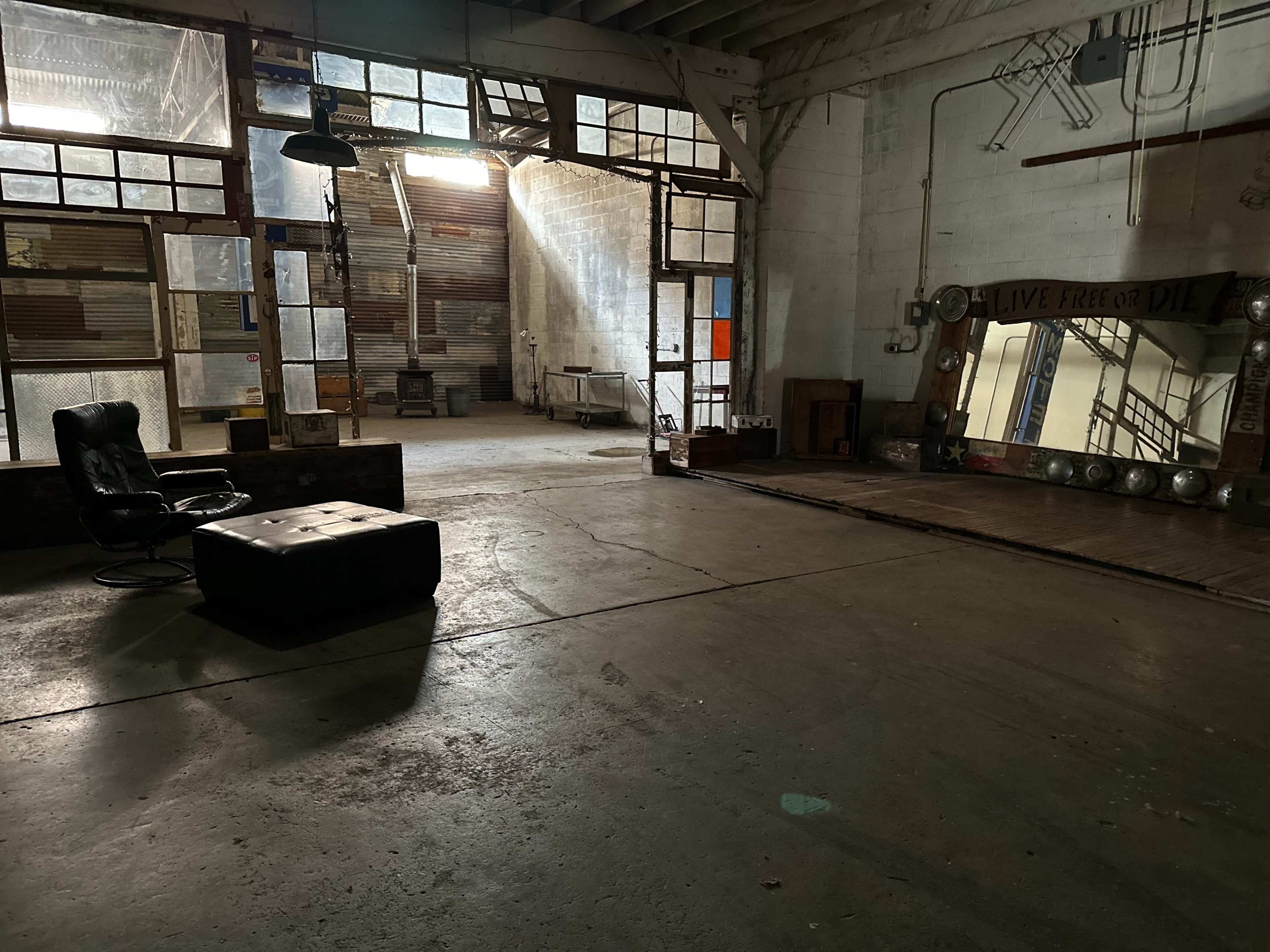 The image shows a spacious, industrial-style room with large windows, a black chair, a mirrored wall, and exposed beams.