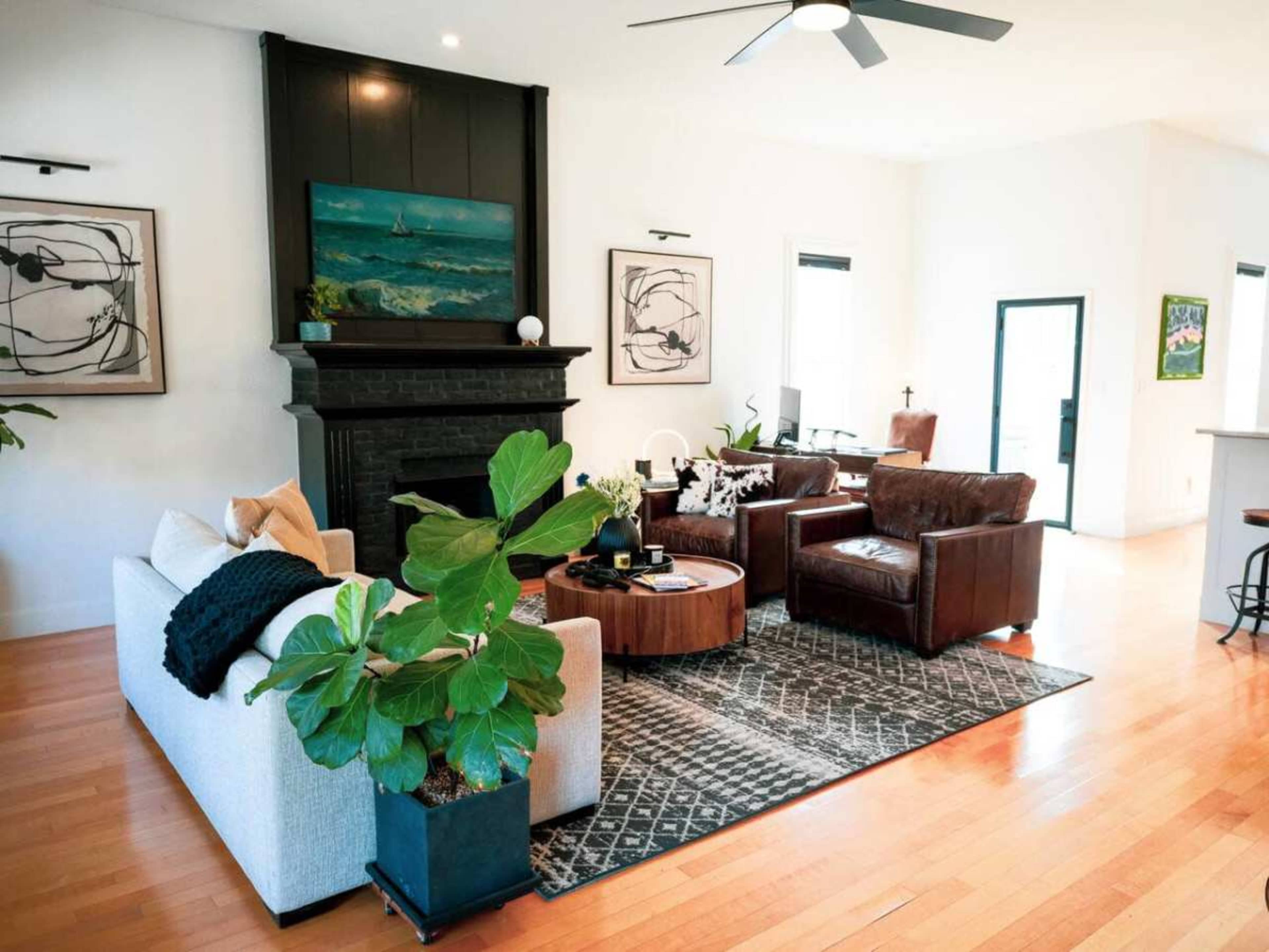 The image shows a modern living room with a white sofa, brown leather armchairs, a round coffee table, and a green plant, all arranged around a patterned rug and a black fireplace.