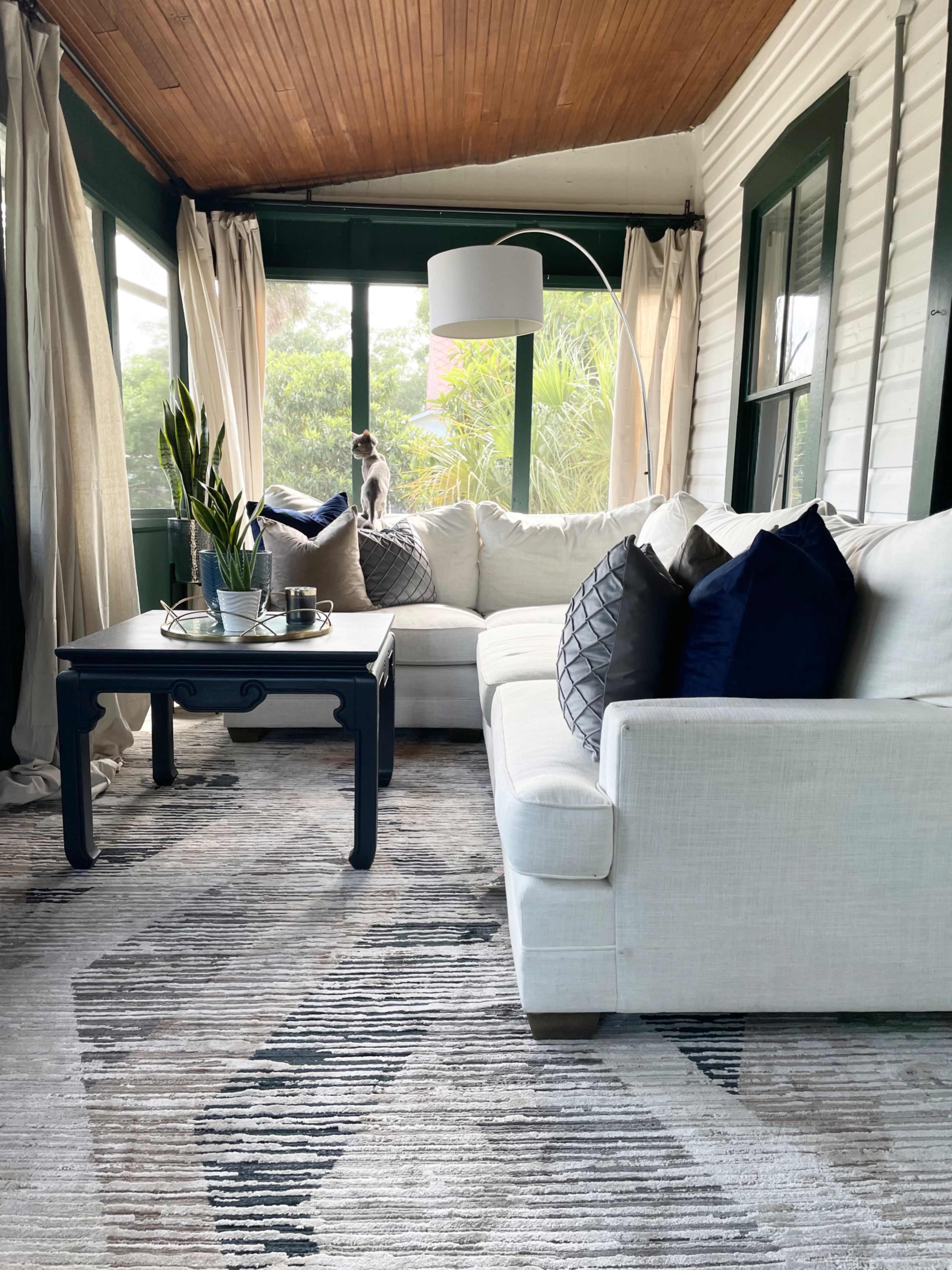 A bright sunroom features a white sectional sofa with navy accent pillows, a dark table in front, and a decorative plant by a window, with a small dog sitting on the couch.