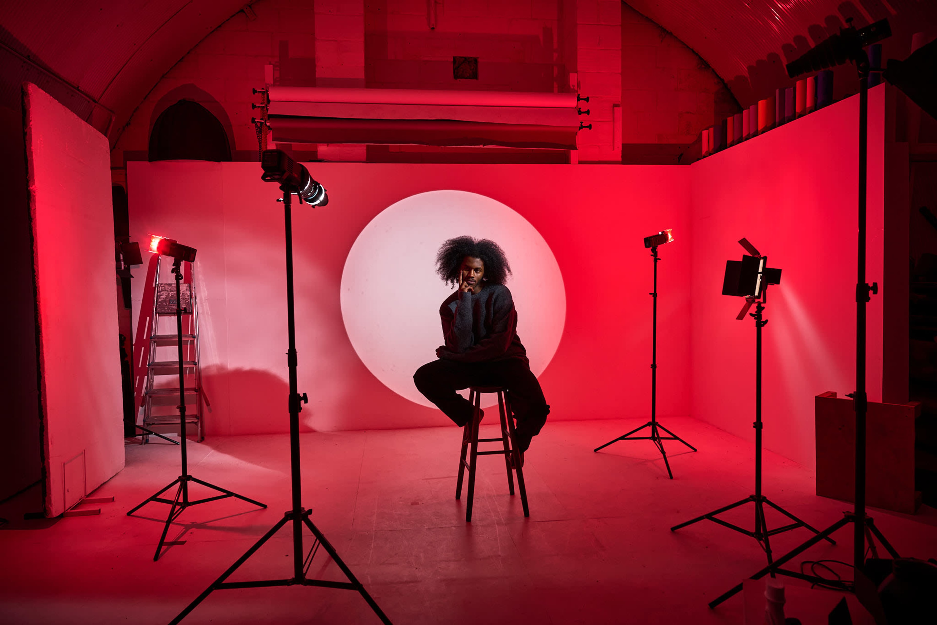 A person with styled hair sits on a stool in front of a large illuminated circular backdrop surrounded by studio lights.