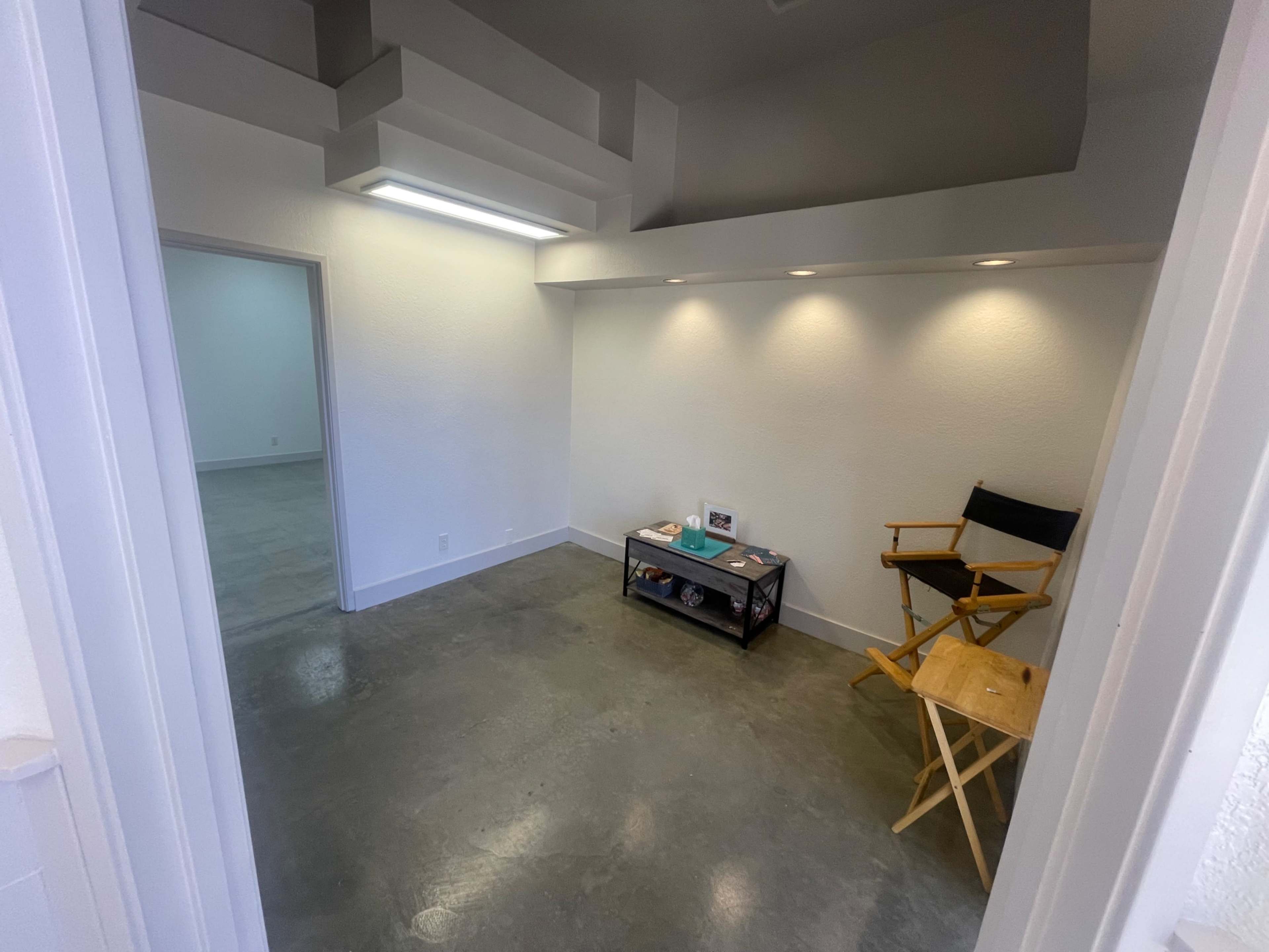 The image shows a small, empty room with a concrete floor, a wooden chair, and a low table against one wall, illuminated by overhead lights.
