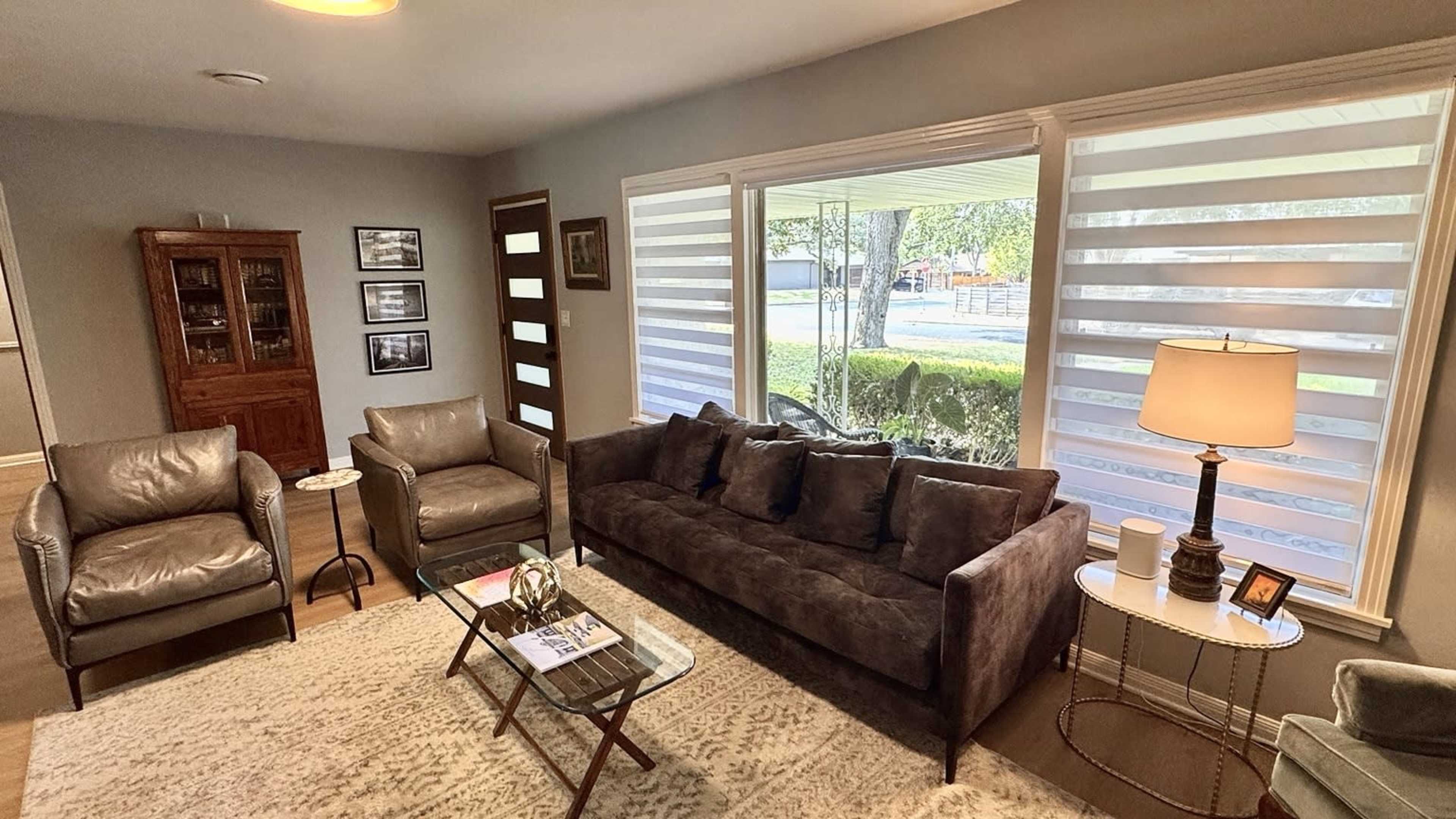 A cozy living room features a dark sofa, two armchairs, a coffee table, and large windows with layered shades allowing natural light to filter in.