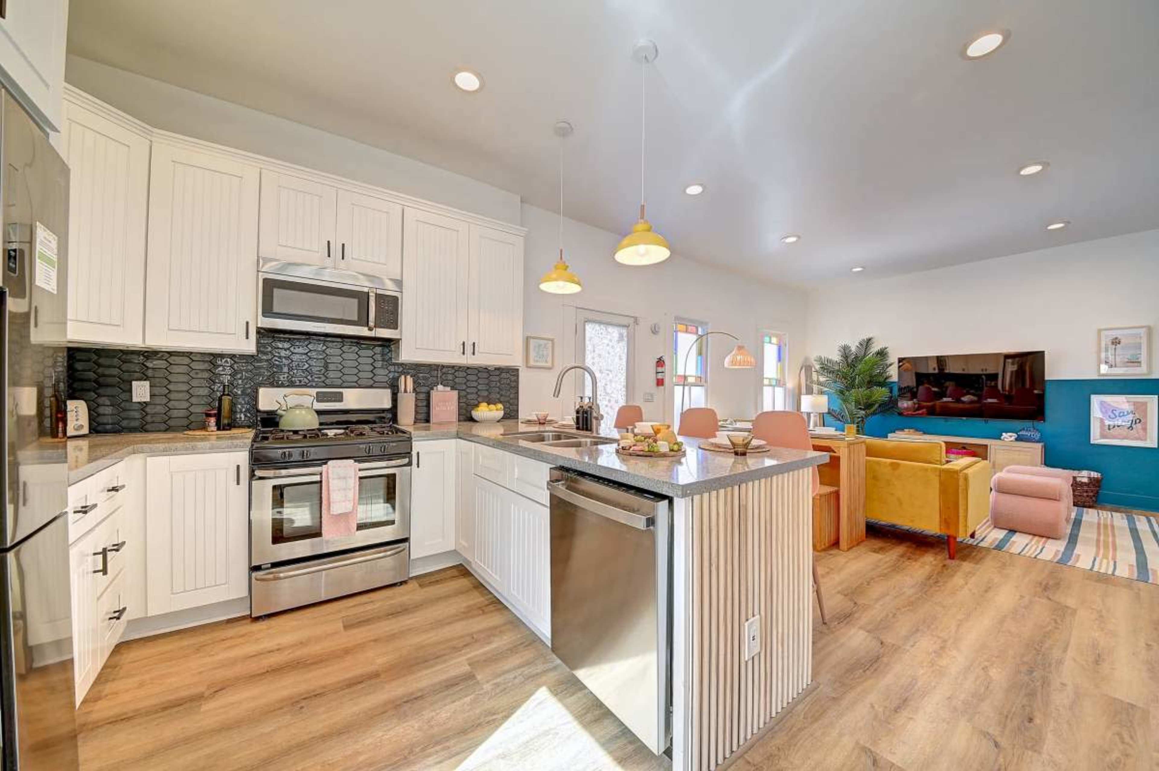 The image shows a modern kitchen with white cabinets, stainless steel appliances, and a large island, adjacent to a colorful living area featuring a green sofa and pink chairs.