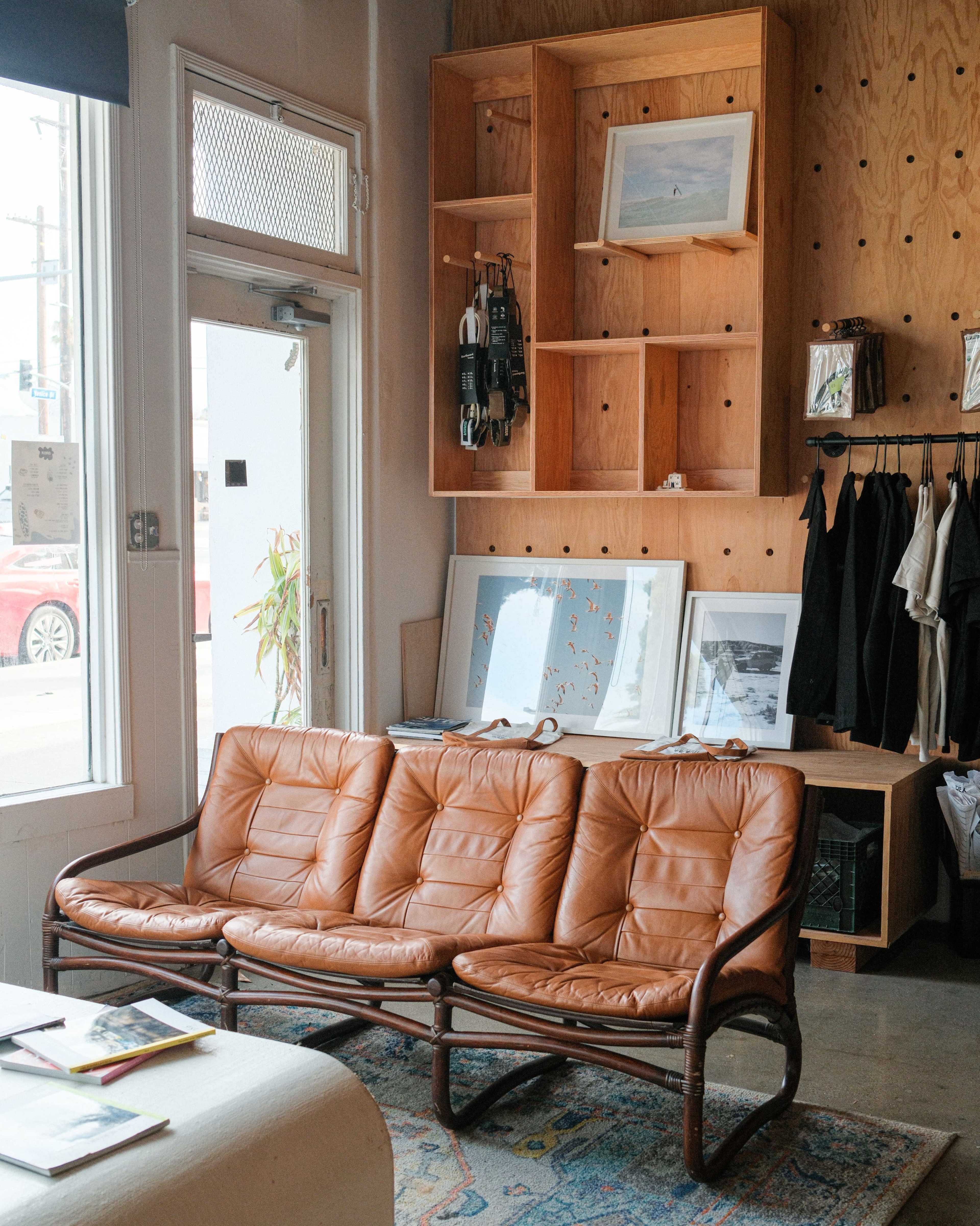 The image features a modern interior with a brown leather sofa, wooden shelves displaying art and decor, and a large window allowing natural light to enter the room.