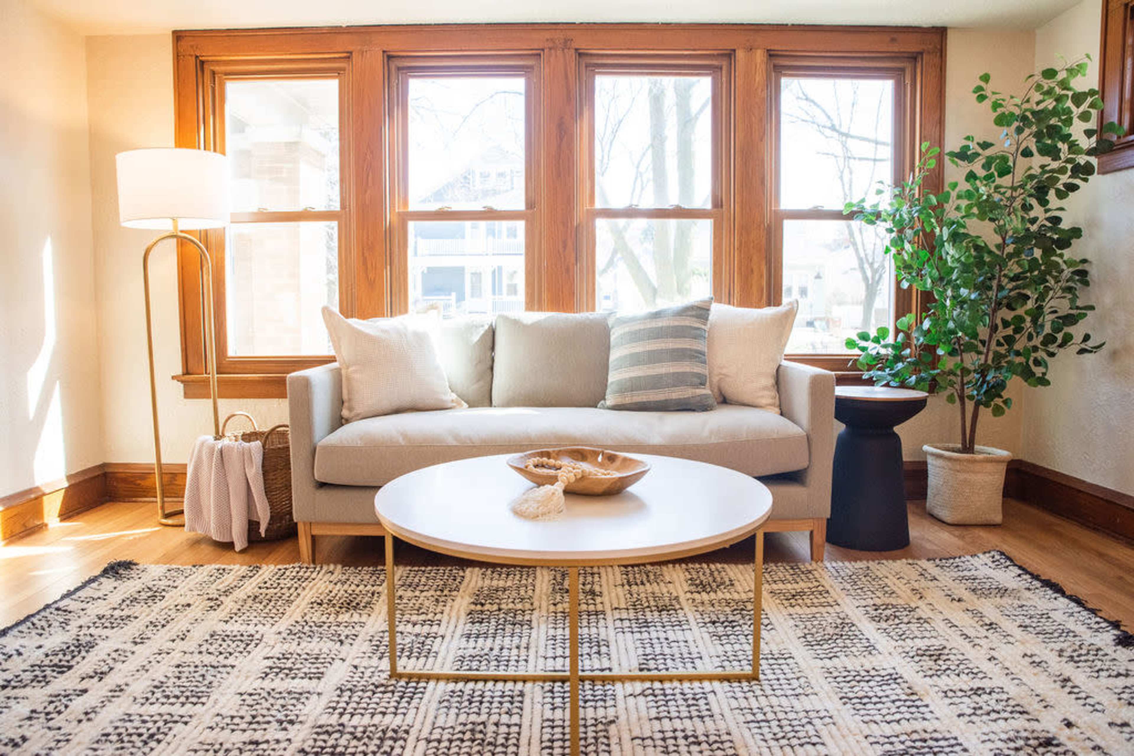 A bright living room features a light-colored sofa with decorative pillows, a round coffee table, and large windows allowing natural light to stream in.