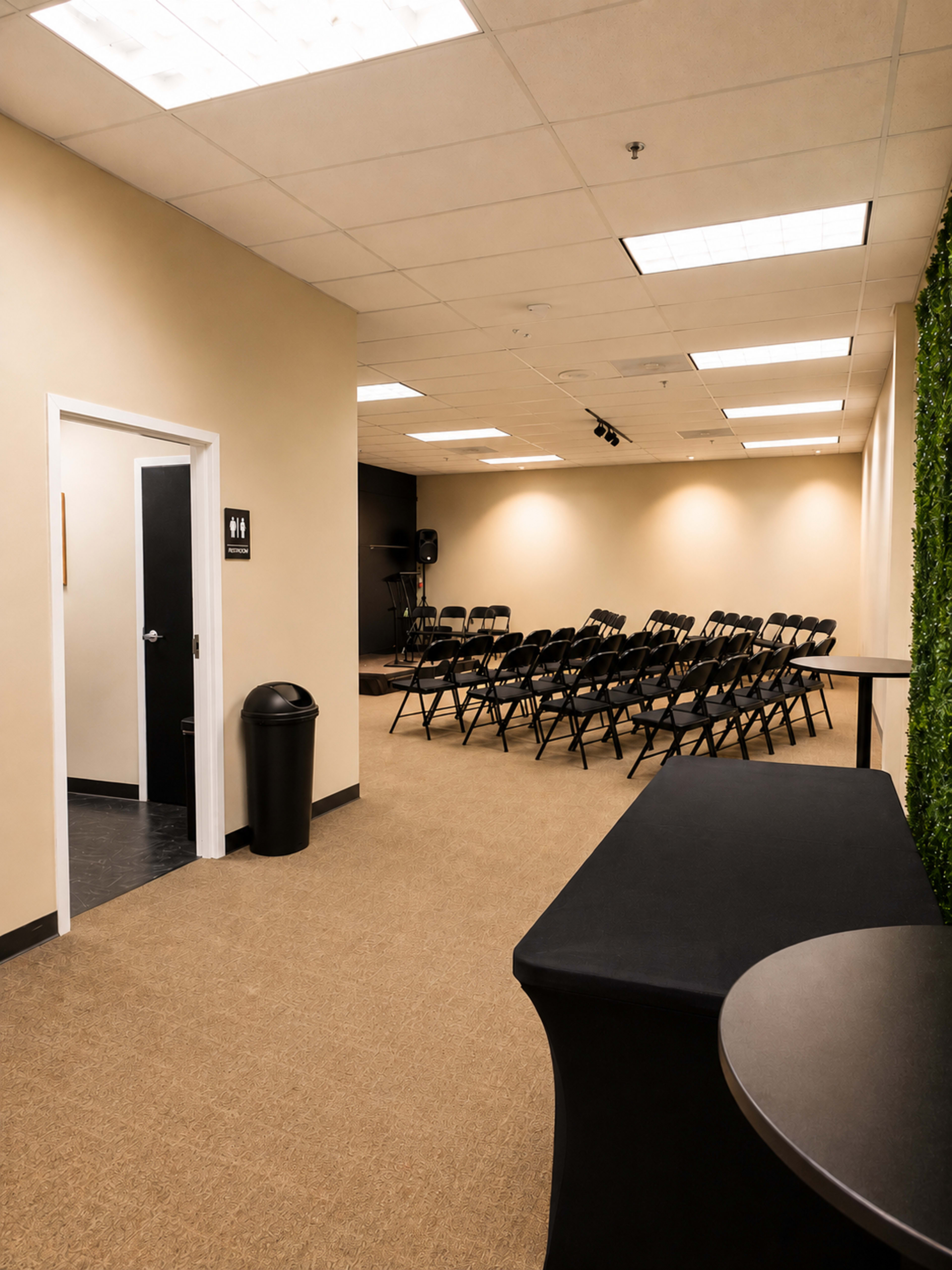 The image shows an empty conference room with rows of black chairs facing a small stage, a trash can in the corner, and a door leading to restrooms on the left.