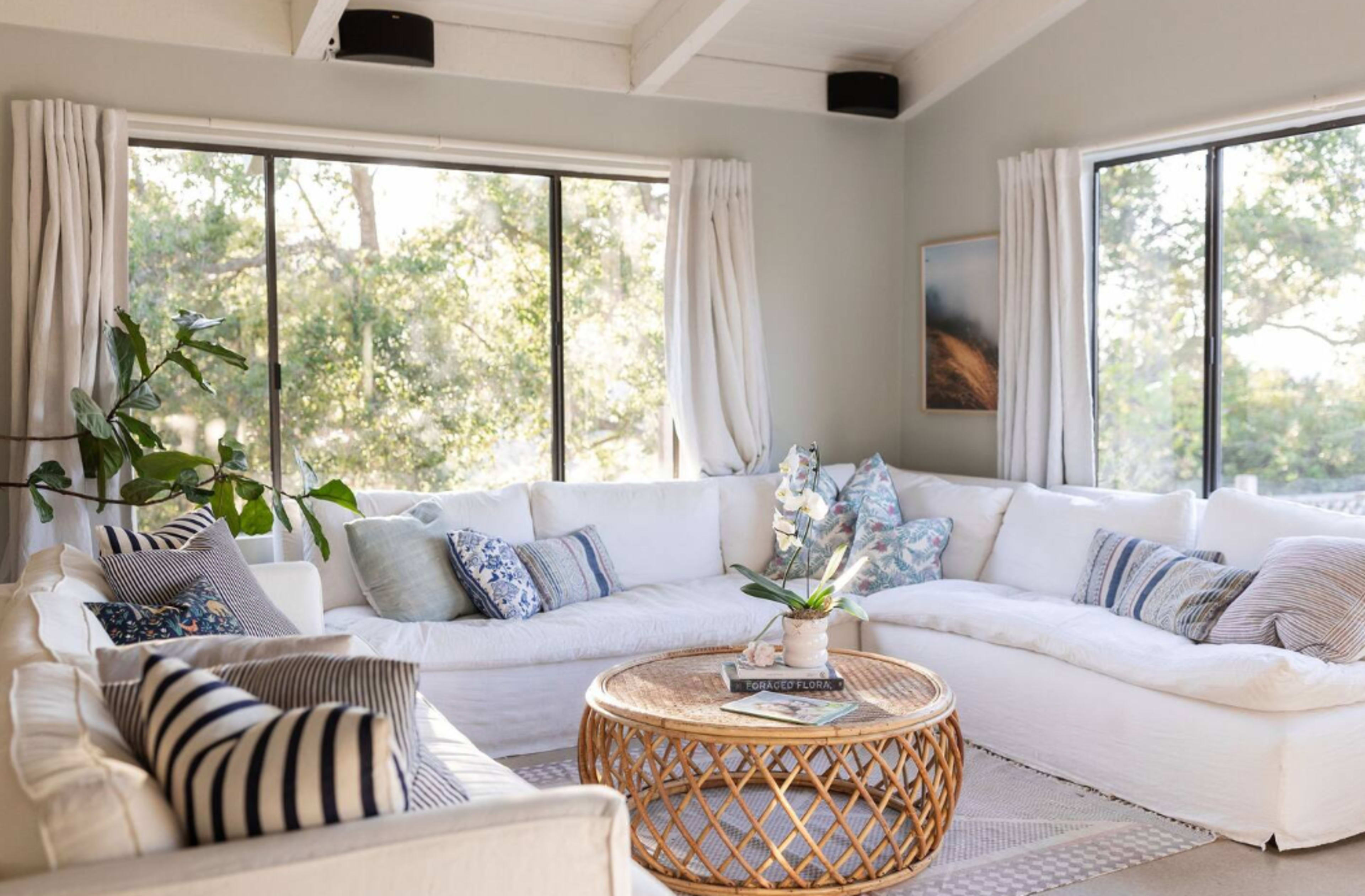 A bright living room with large windows, a white sectional sofa, a round rattan coffee table, and decorative pillows.