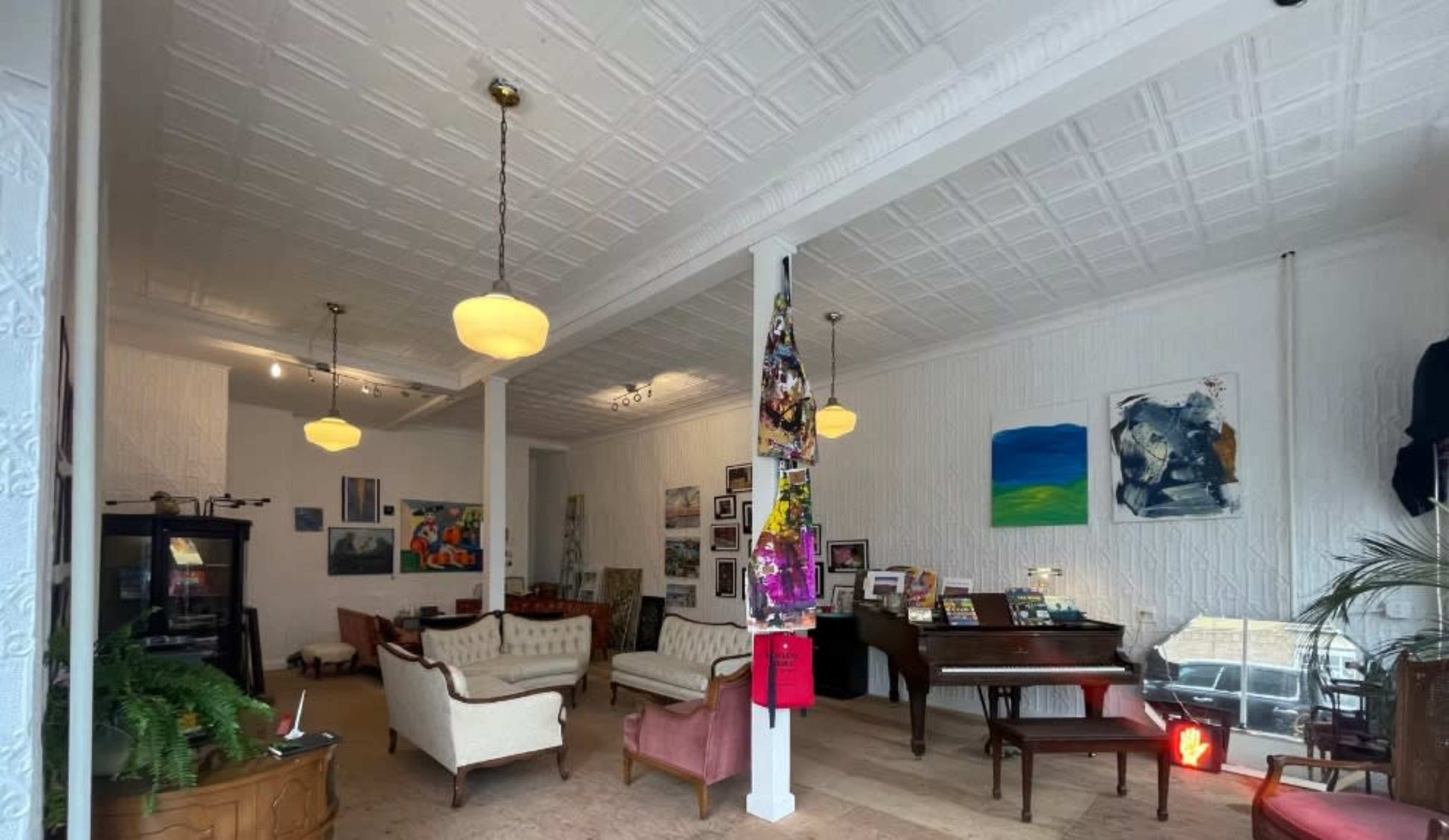 The image shows an interior of a cozy art space featuring a seating area with sofas, a piano, and framed artworks on the walls.