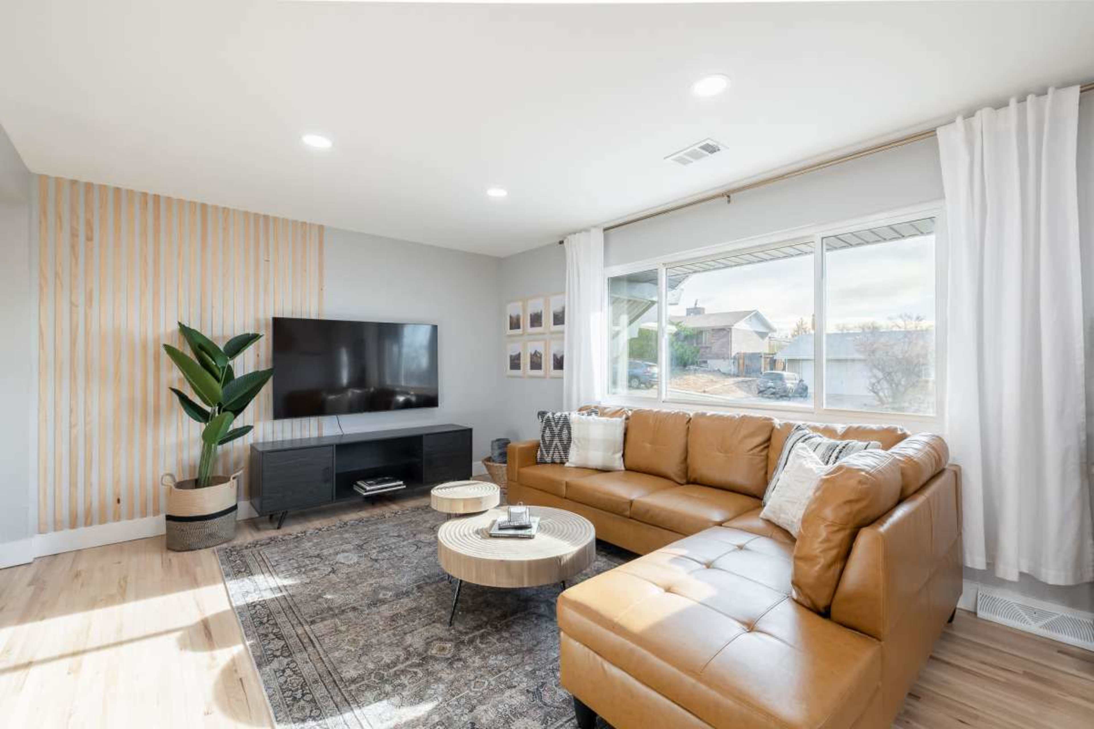 The image depicts a modern living room with a brown leather sectional sofa, a round coffee table, a wall-mounted television, and large windows allowing natural light to enter.
