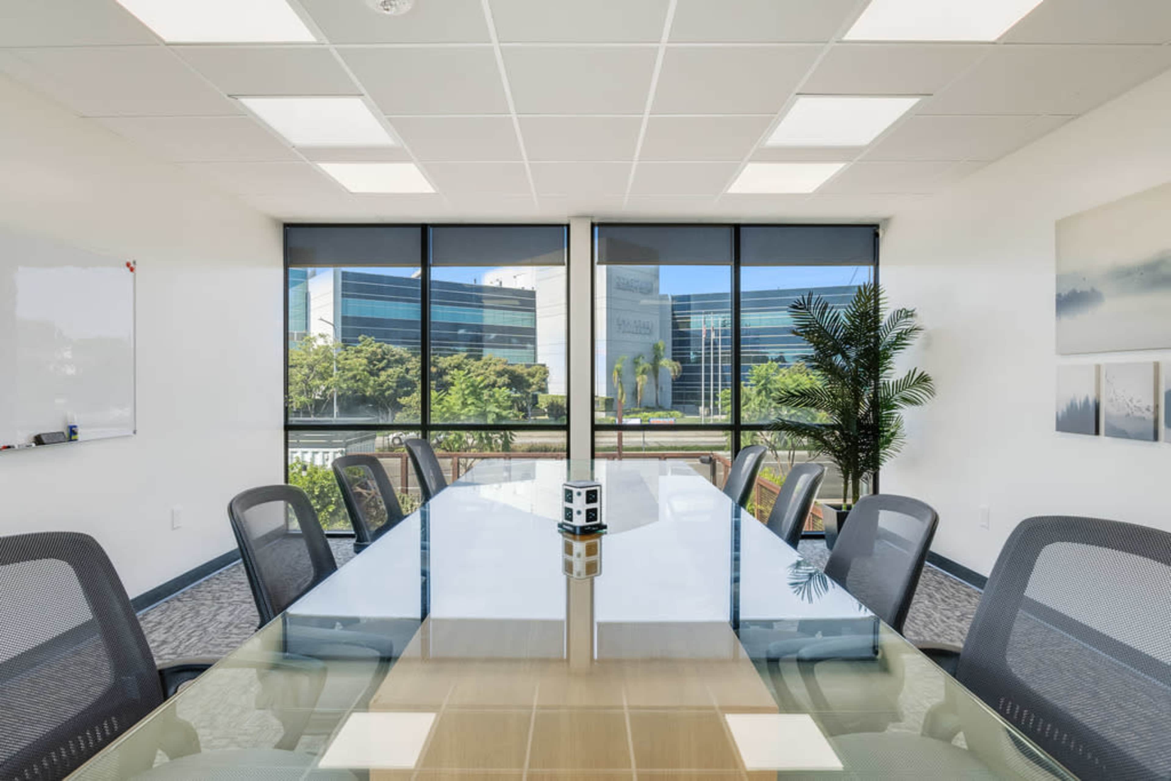 A spacious conference room features a long glass table surrounded by mesh chairs, with large windows offering a view of greenery outside.