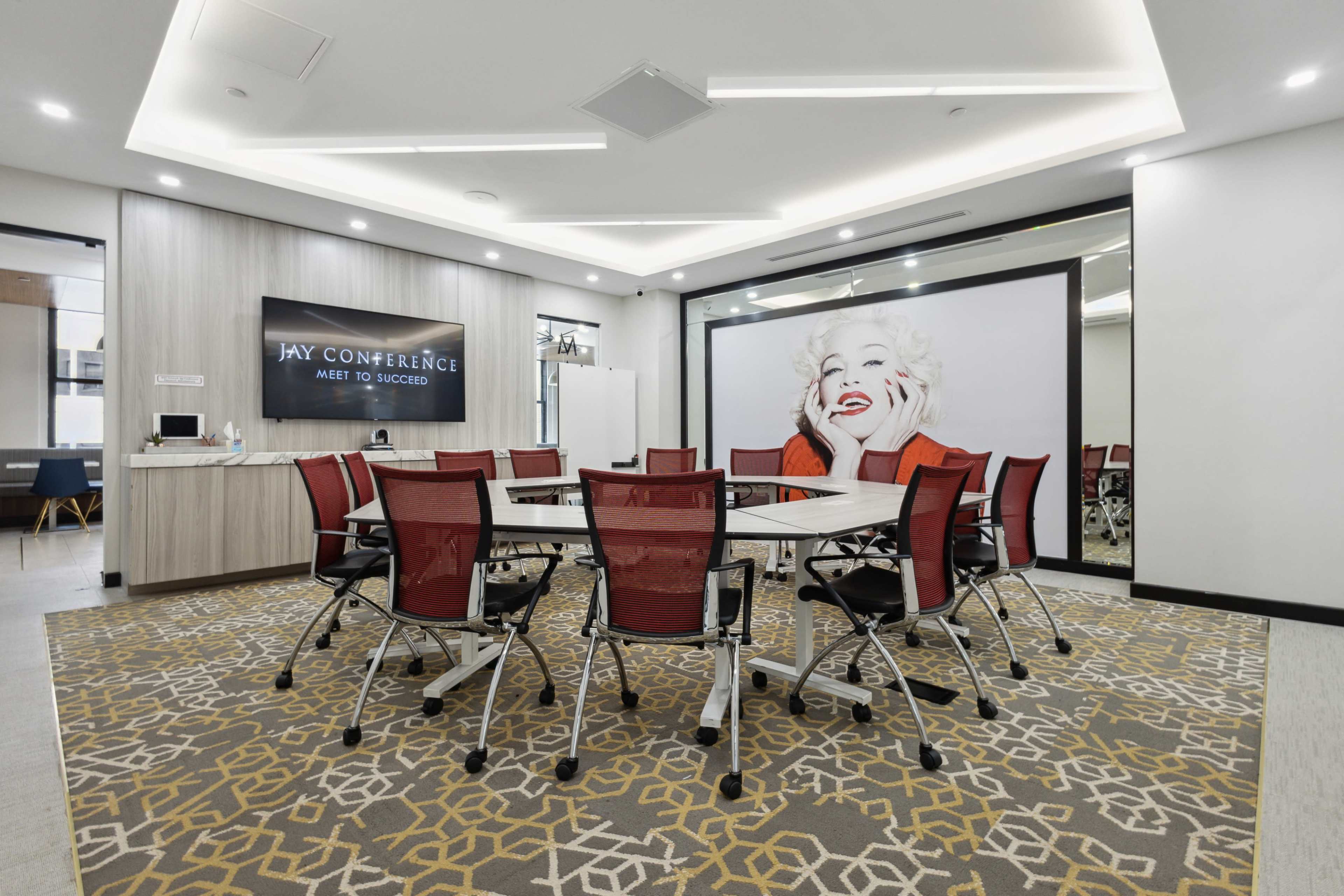 A modern conference room features a large table surrounded by red chairs and a prominent wall mural of a woman.
