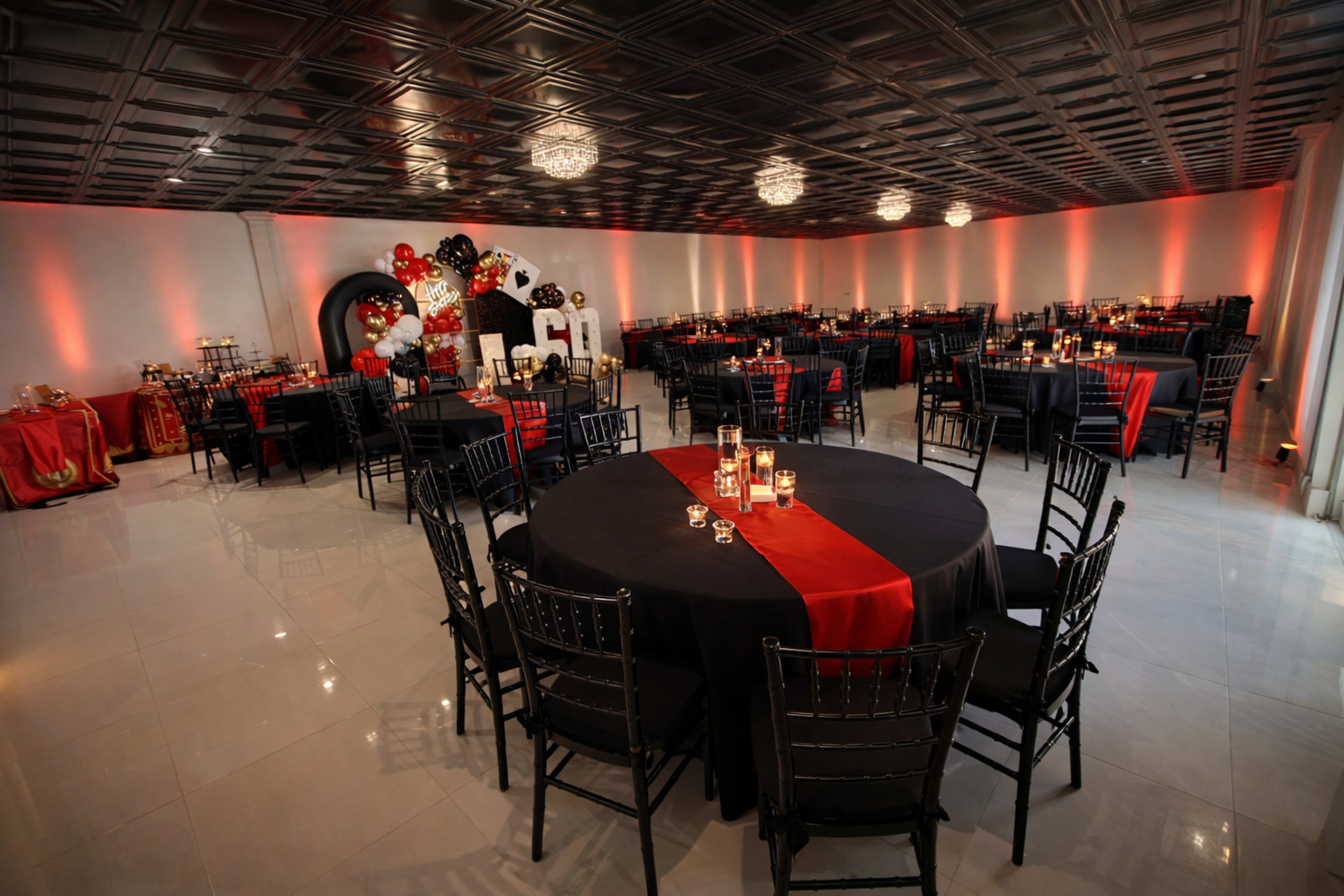 A spacious event room is arranged with black and red tables, each featuring candle centerpieces, under a patterned ceiling and ambient lighting.