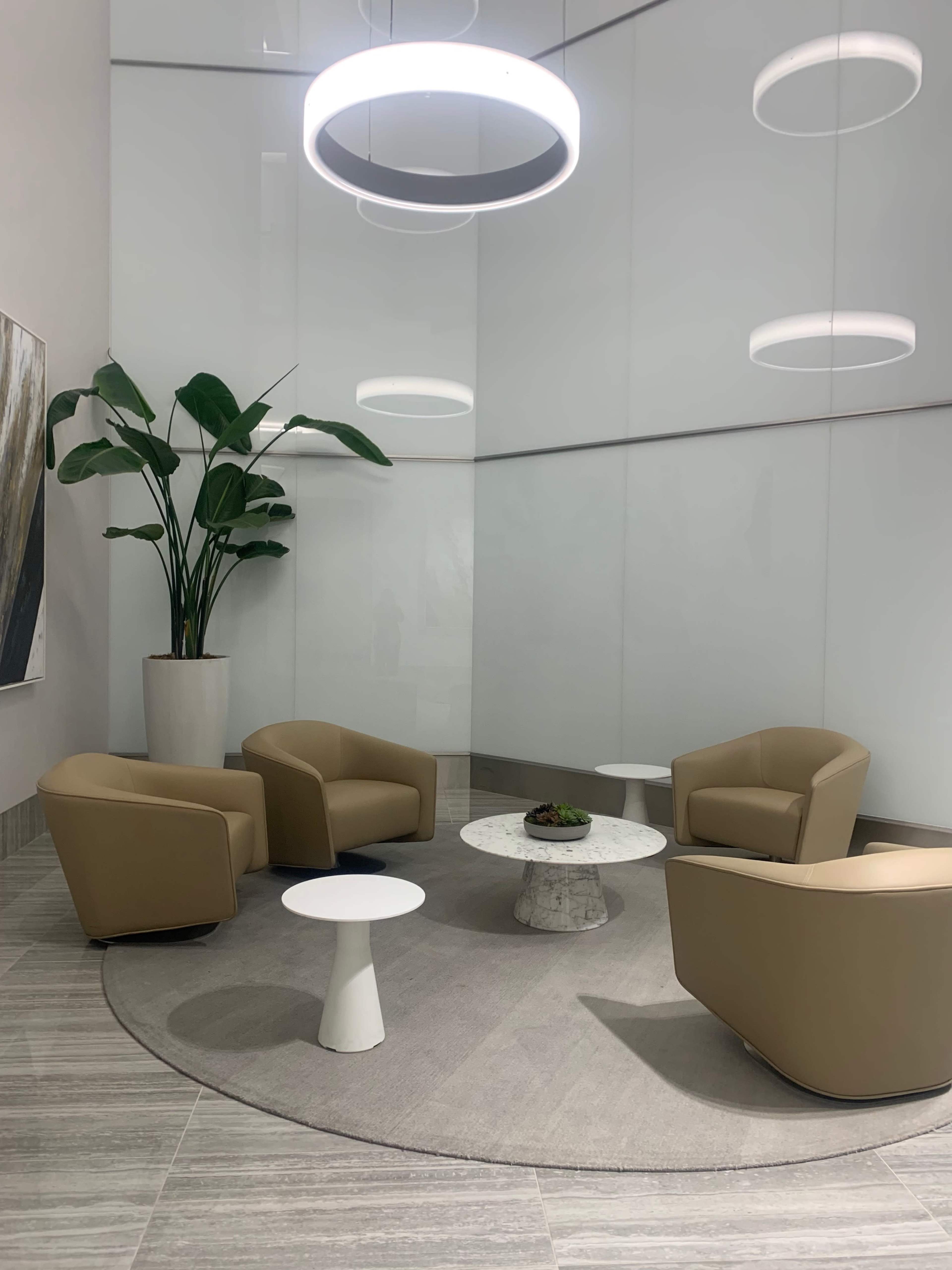 A modern seating area with two beige armchairs, a white round coffee table, and a large plant in a corner, all set against a sleek, minimalist background.